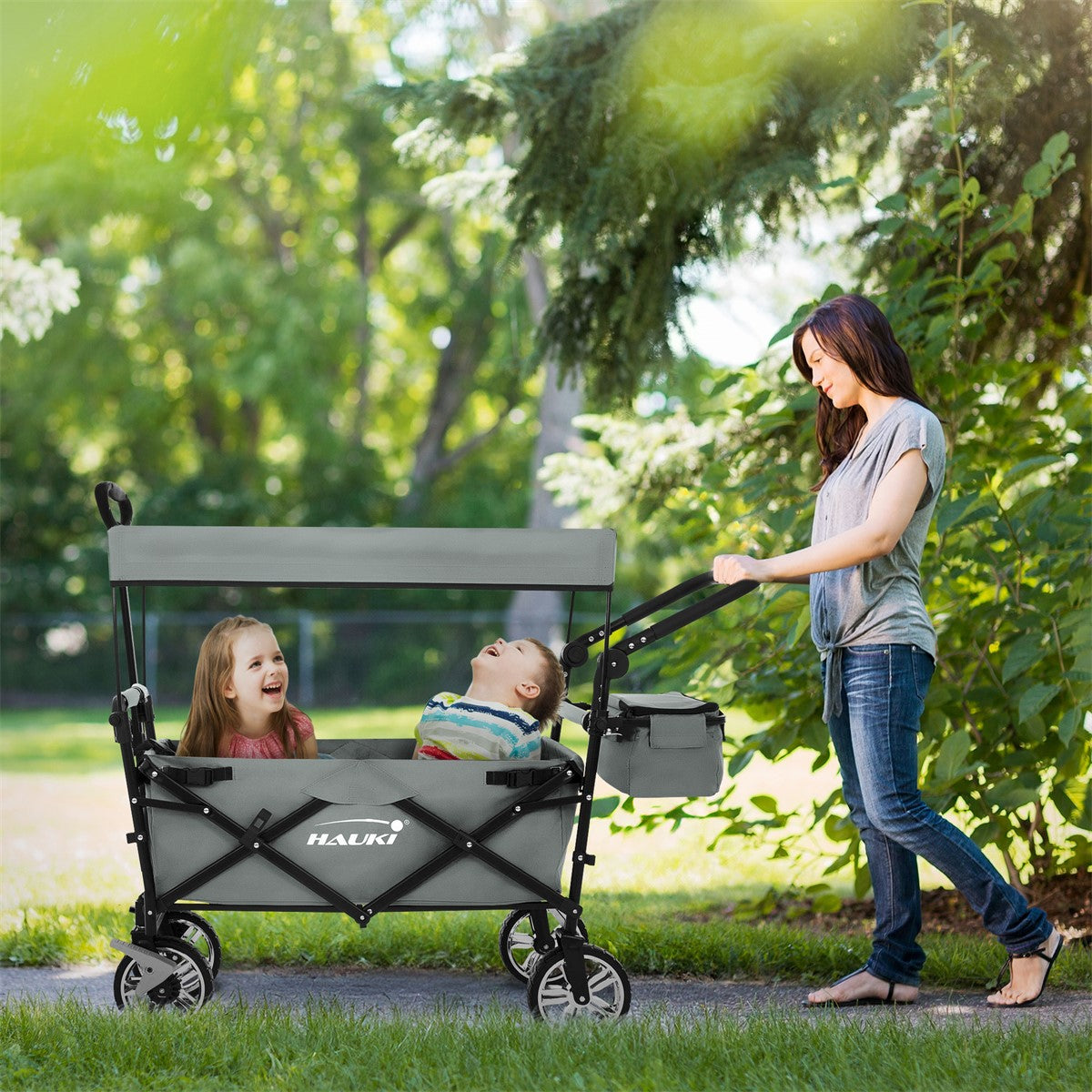 Eine Frau schiebt einen grauen Kinderwagen mit zwei Kindern im Park.