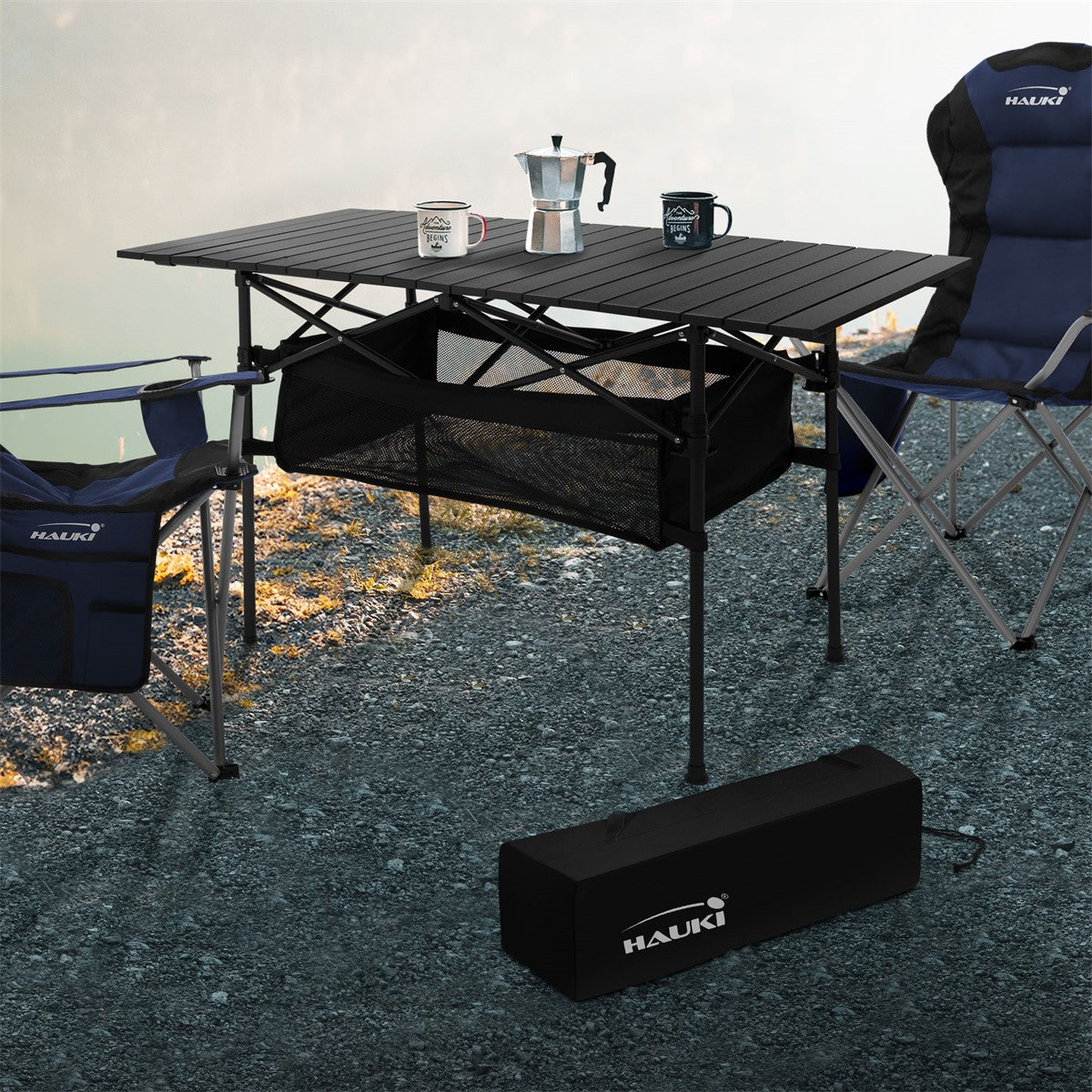 Table with a coffee pot and two chairs on a gravel path by the water.