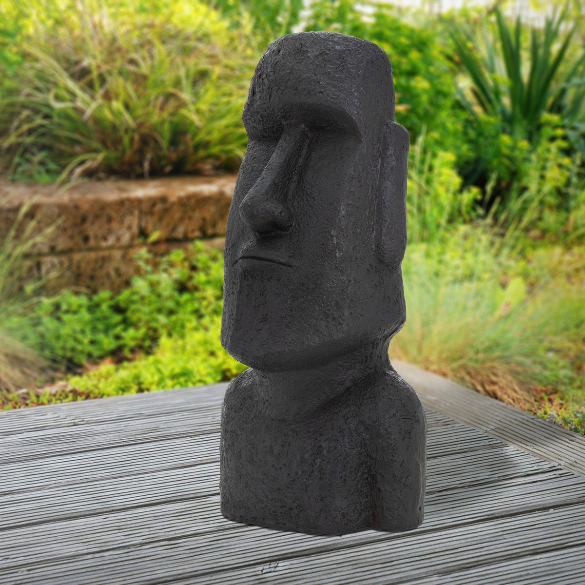 Schwarze Moai-Statue auf einem Holzuntergrund, umgeben von grüner Vegetation.