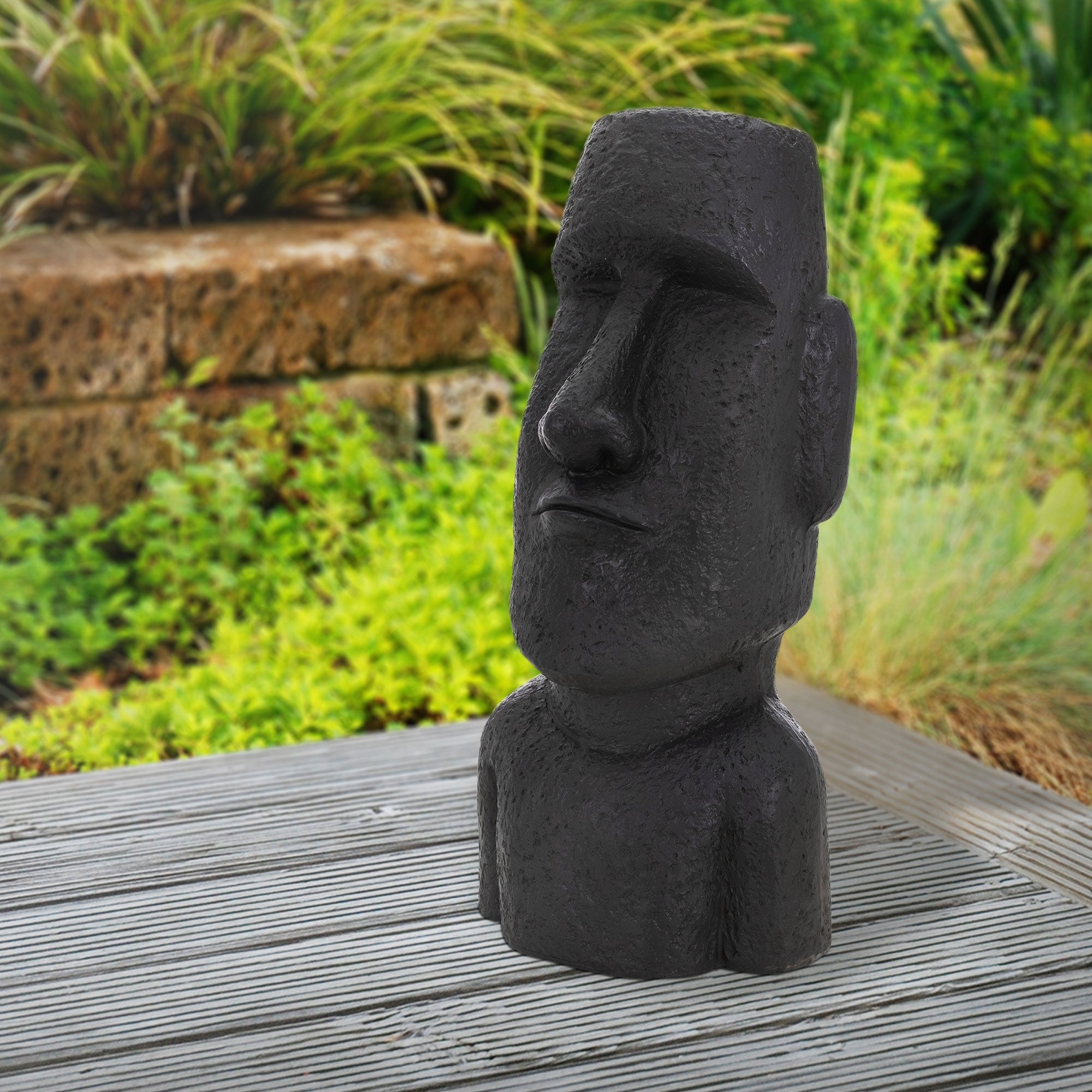 Schwarze Statue eines Moai auf einem Holztisch, im Hintergrund grüne Pflanzen.