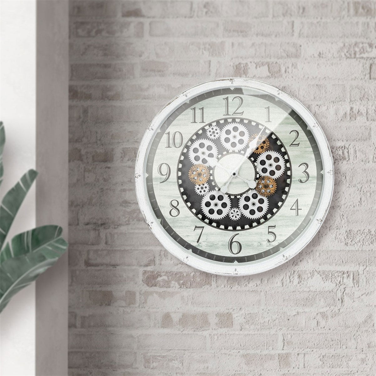 Ronde wandklok met tandwielen aan een lichte muur en een plant op de voorgrond.