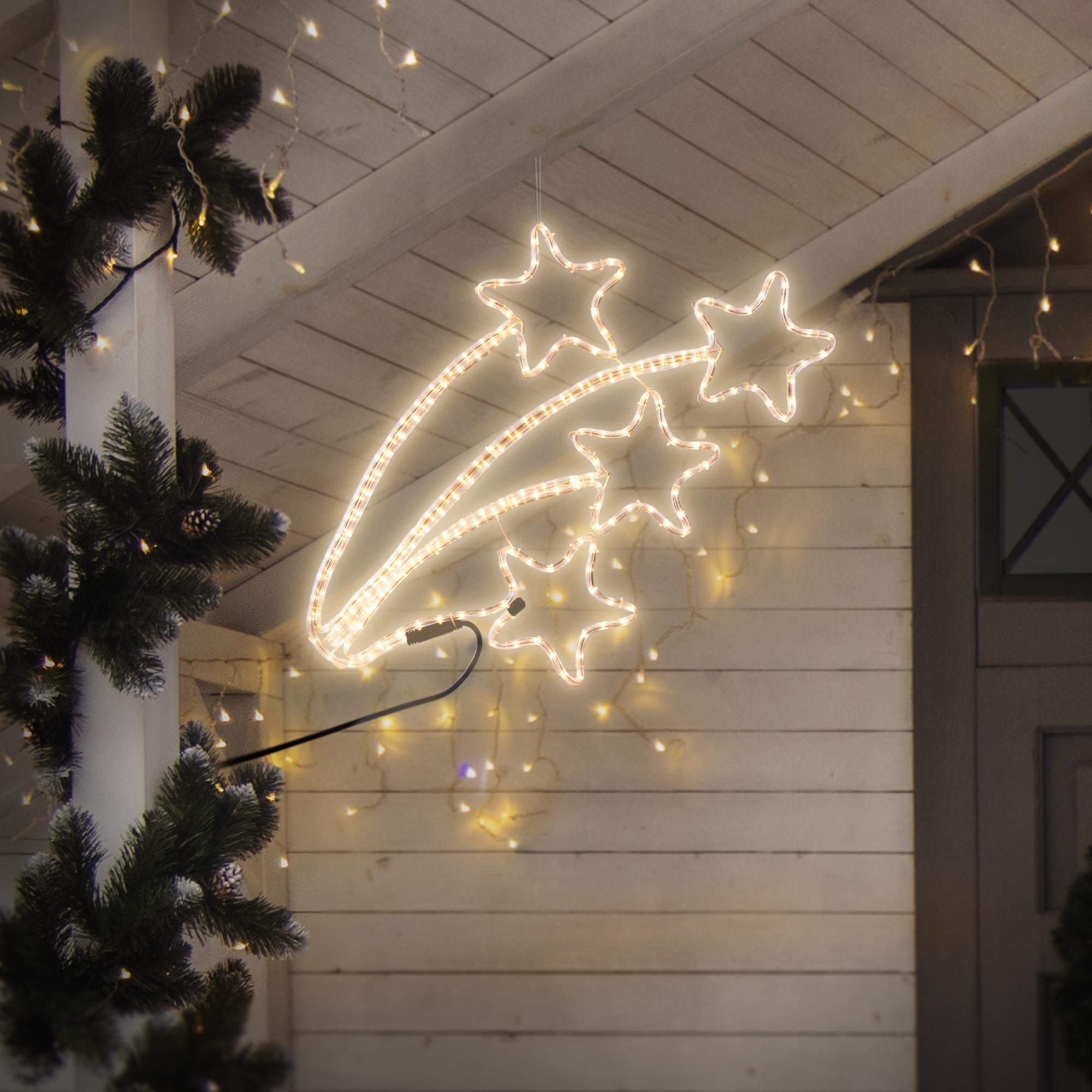 Étoiles lumineuses faites de lumières, suspendues à un toit, entourées de branches de sapin.