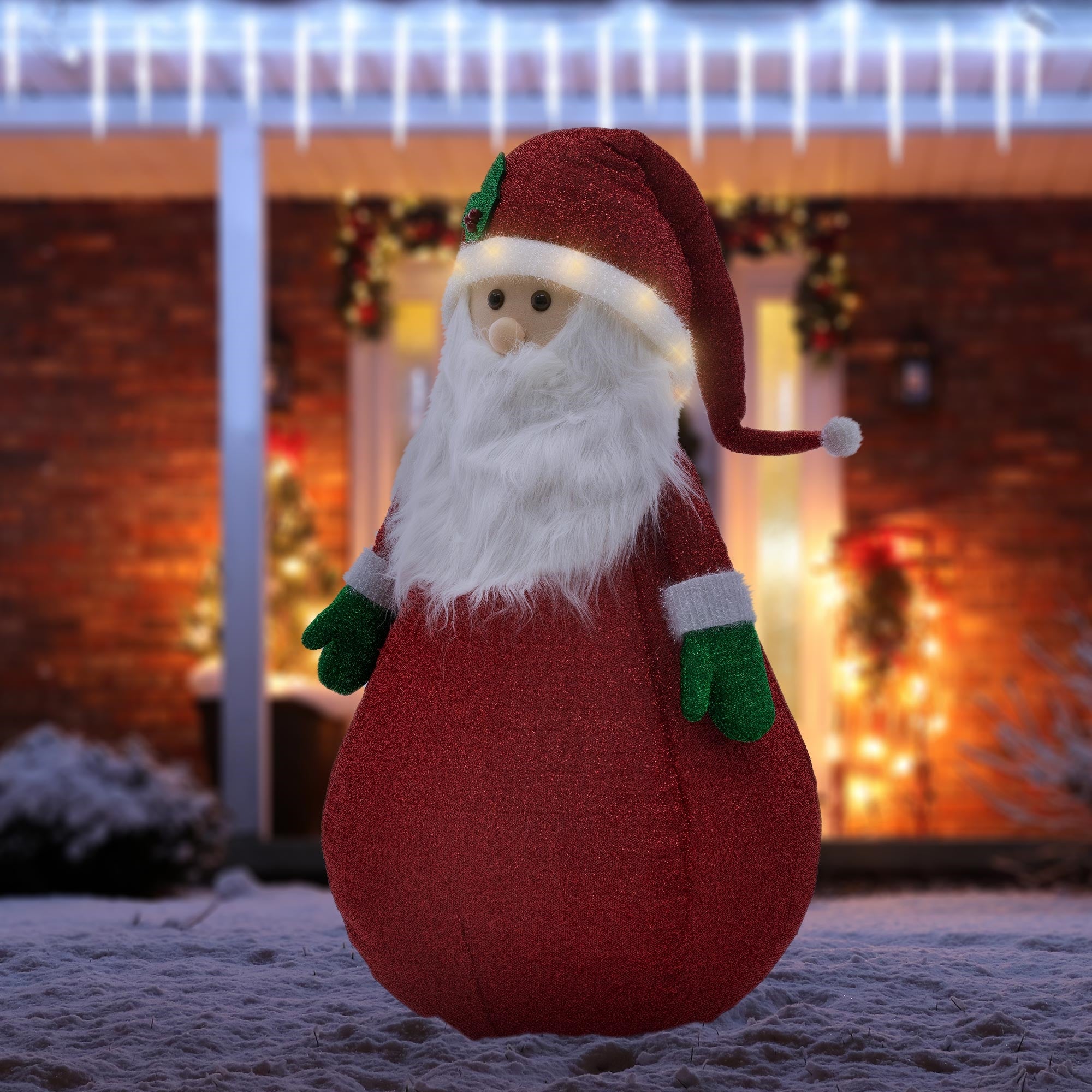 Un Babbo Natale di stoffa sta nella neve davanti a una casa illuminata.
