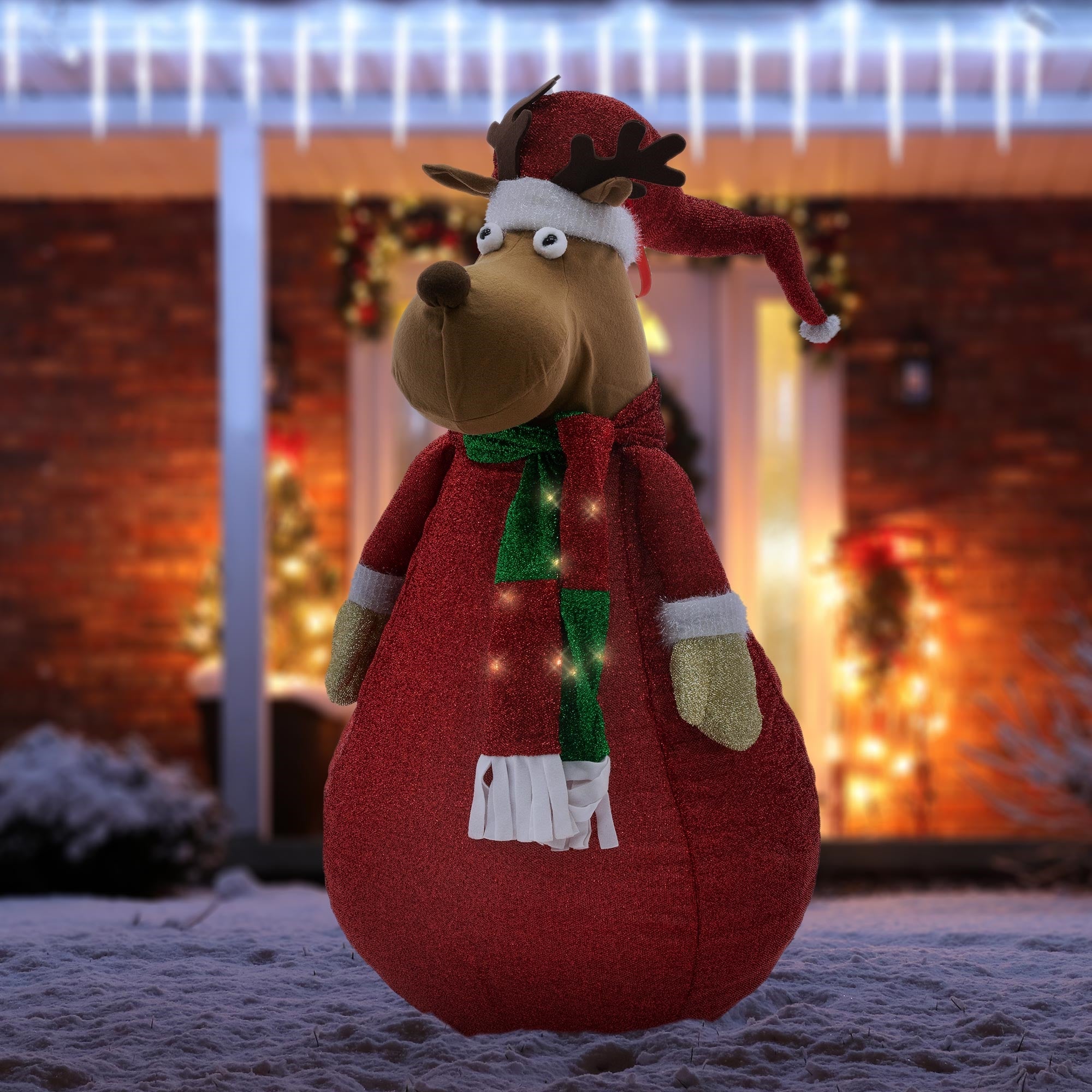 Plüschrentier in Weihnachtsdekoration im Schnee, mit Lichtern im Hintergrund.