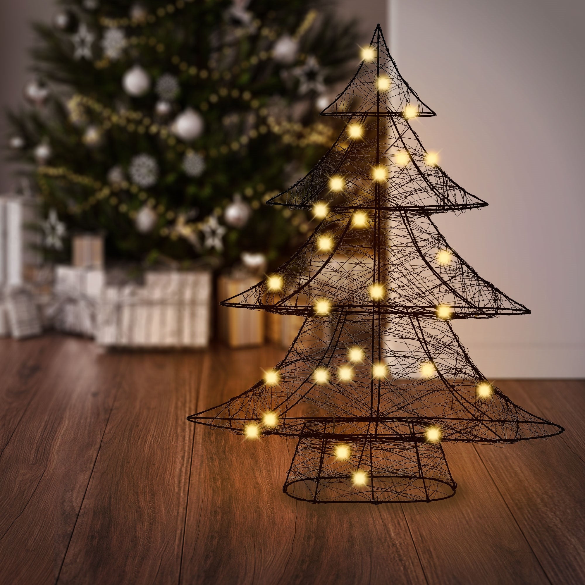 Schwarzer Drahtbaum mit Lichtern vor einem Weihnachtsbaum und Geschenken.