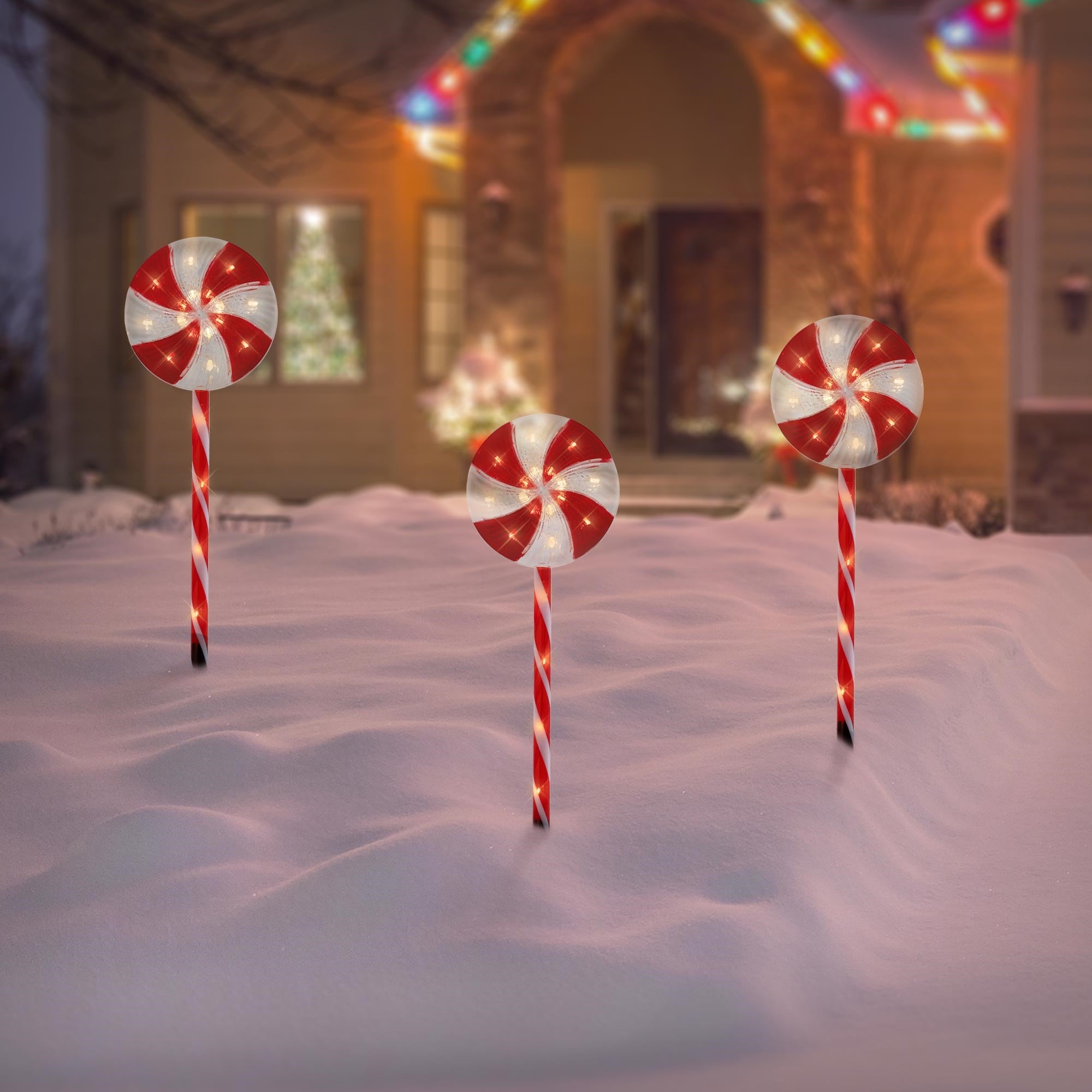 Tres grandes piruletas brillantes están en la nieve frente a una casa decorada festivamente.