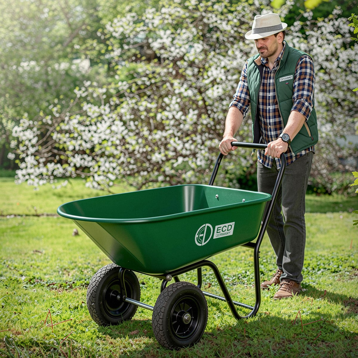 Ein Mann schiebt eine grüne Schubkarre über eine Wiese mit blühenden Bäumen.