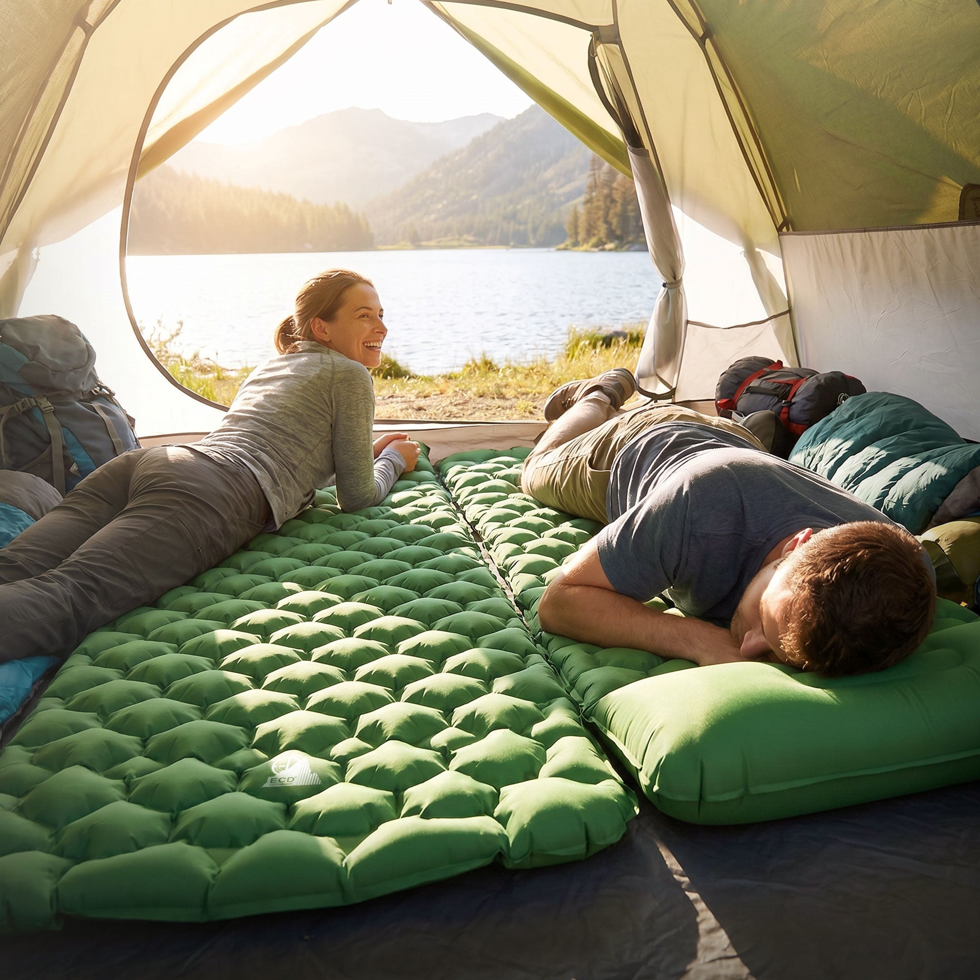 Two people lying in a tent on sleeping mats, view of a lake and mountains in the background.