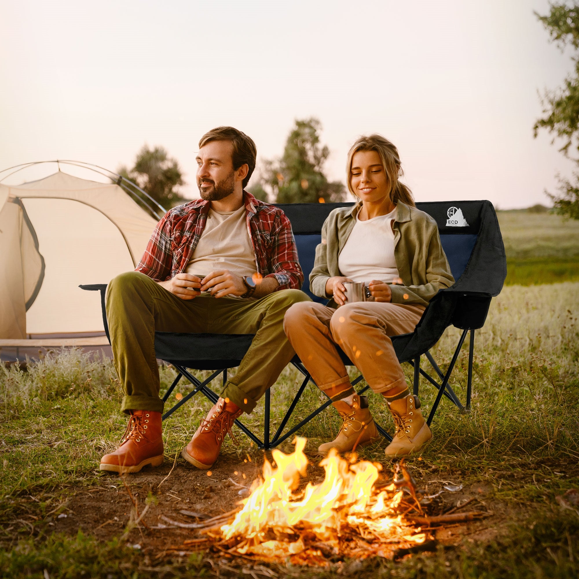 Paar entspannt in Campingstühlen vor einem Zelt, mit einem Feuer in der Mitte.