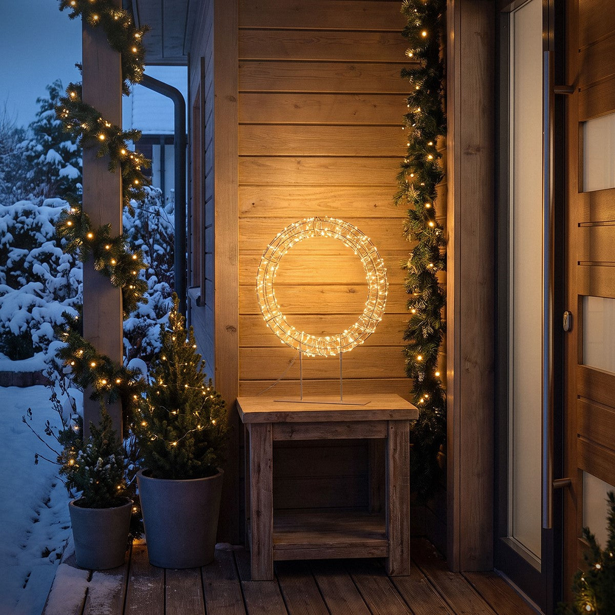 Beleuchteter Kranz auf einem Tisch vor einem Holzhaus, umgeben von Schnee.