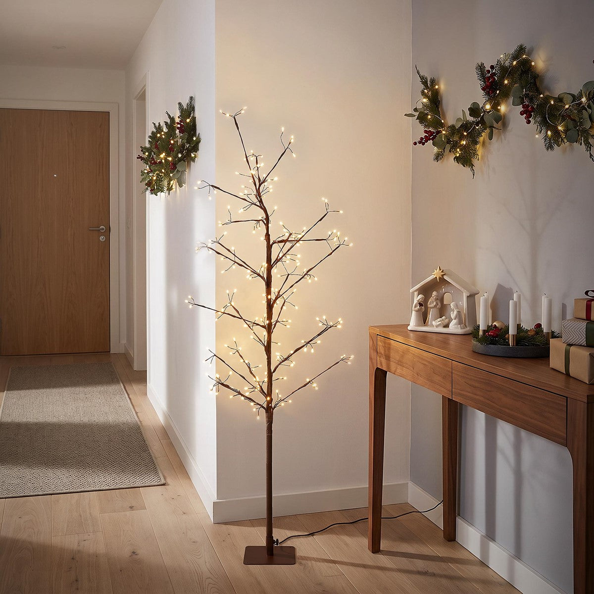 Schlanker, kahler Baum mit leuchtenden Lichterketten auf einem braunen Standfuß in einem Flur neben einer geschmückten Kommode.