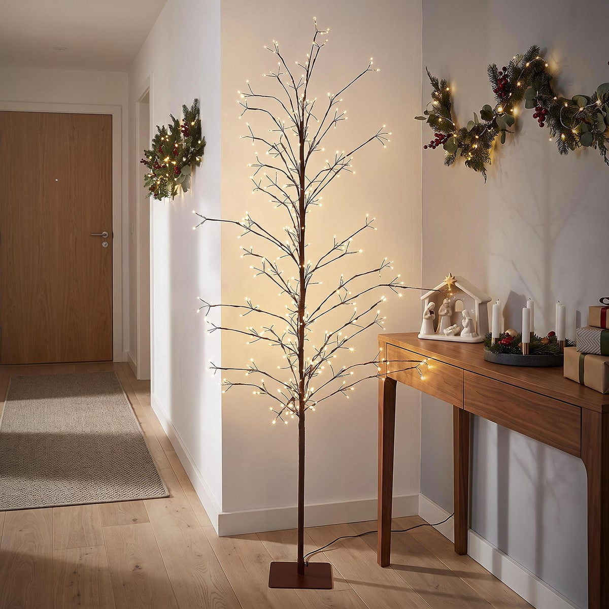 Schlanker, kahler Baum mit leuchtenden Lichterketten auf einem braunen Standfuß in einem Flur neben einer geschmückten Kommode.