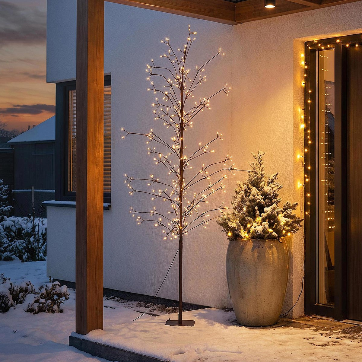 Beleuchteter künstlicher Baum neben einem bepflanzten Topf auf einer verschneiten Veranda bei Dämmerung.