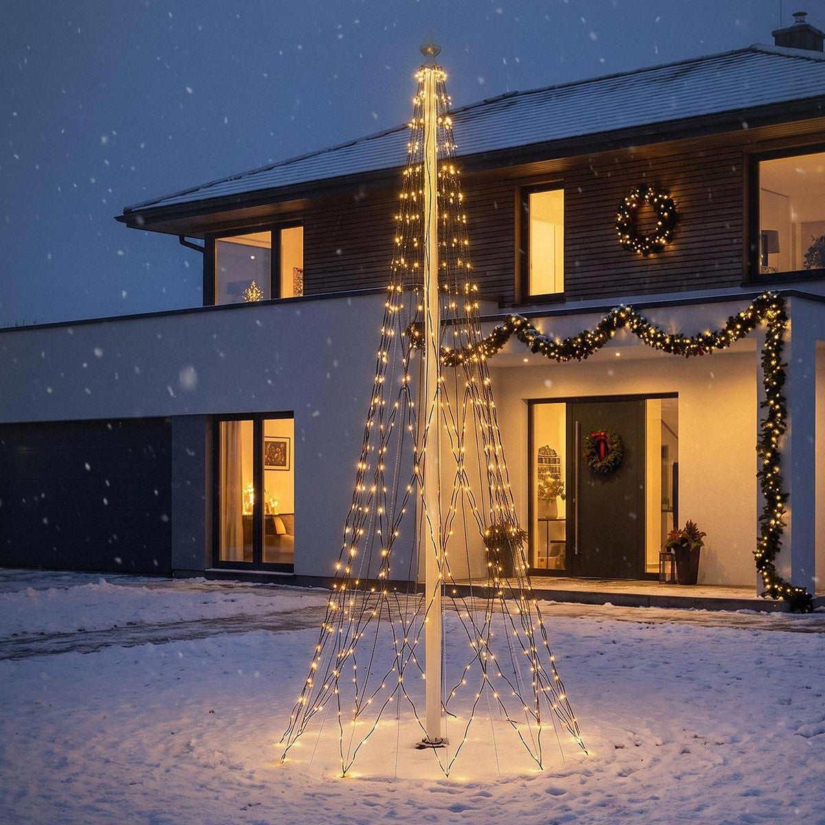 Et oplyst lysttræ står i en snedækket have foran et julepyntet hus.