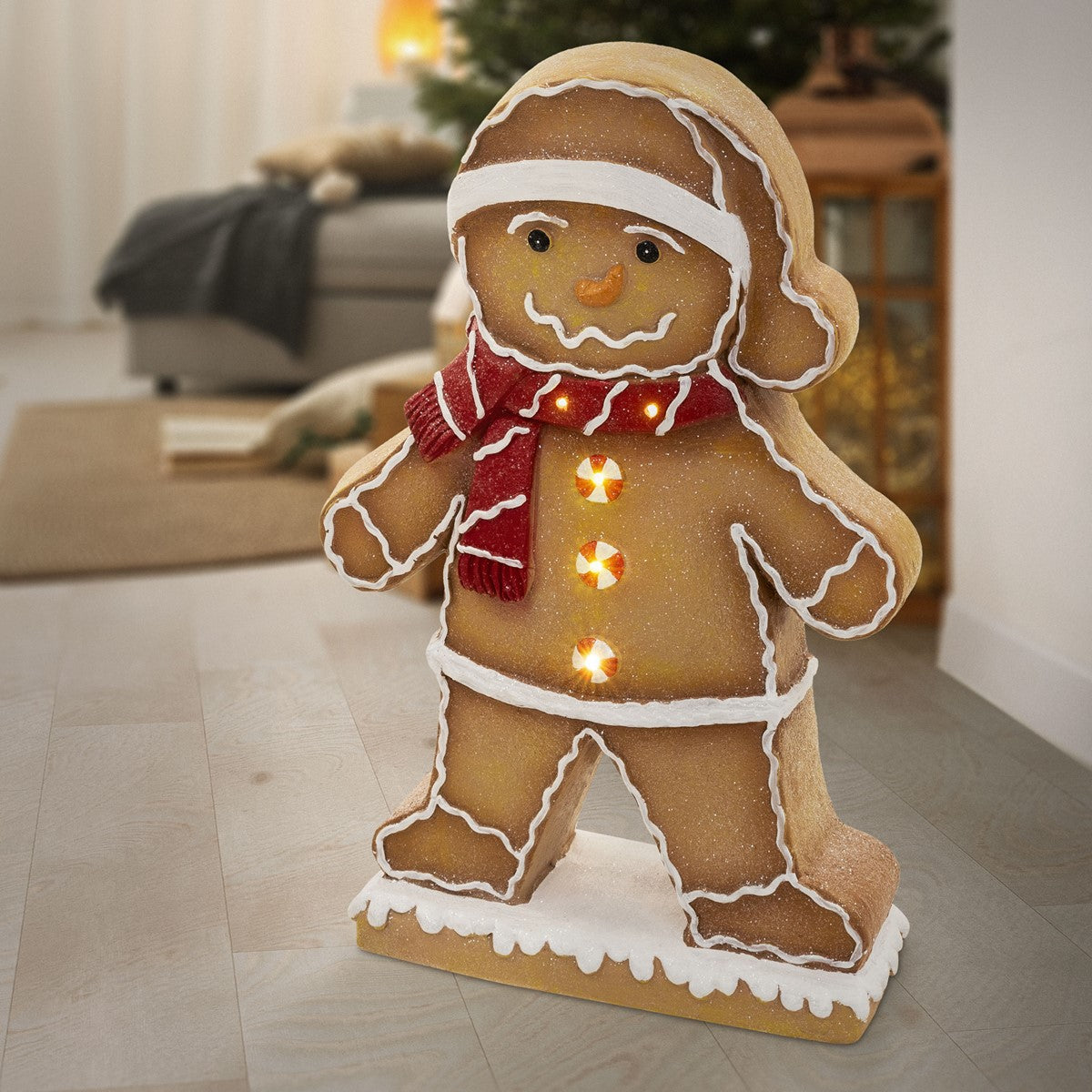 Lebkuchenmann mit rotem Schal und Mütze, beleuchtet, vor einem Weihnachtsbaum.