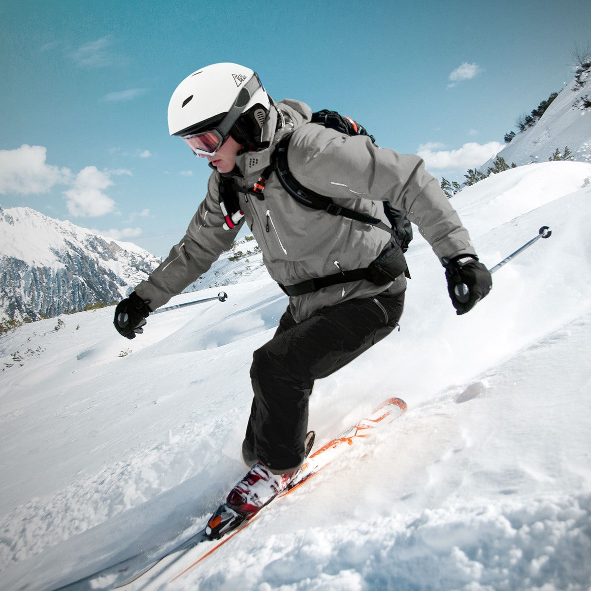 Skifahrer in grauer Jacke fährt auf schneebedecktem Hang mit Bergen im Hintergrund.