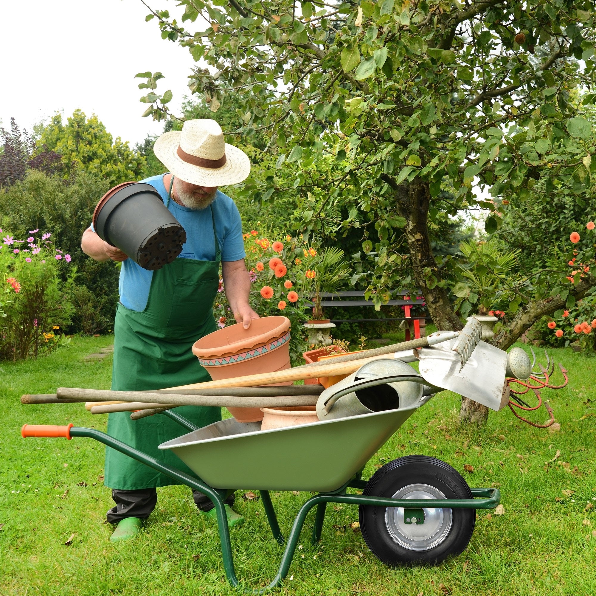 Mann im Garten mit Schubkarre, Töpfen und Werkzeugen, umgeben von Blumen.