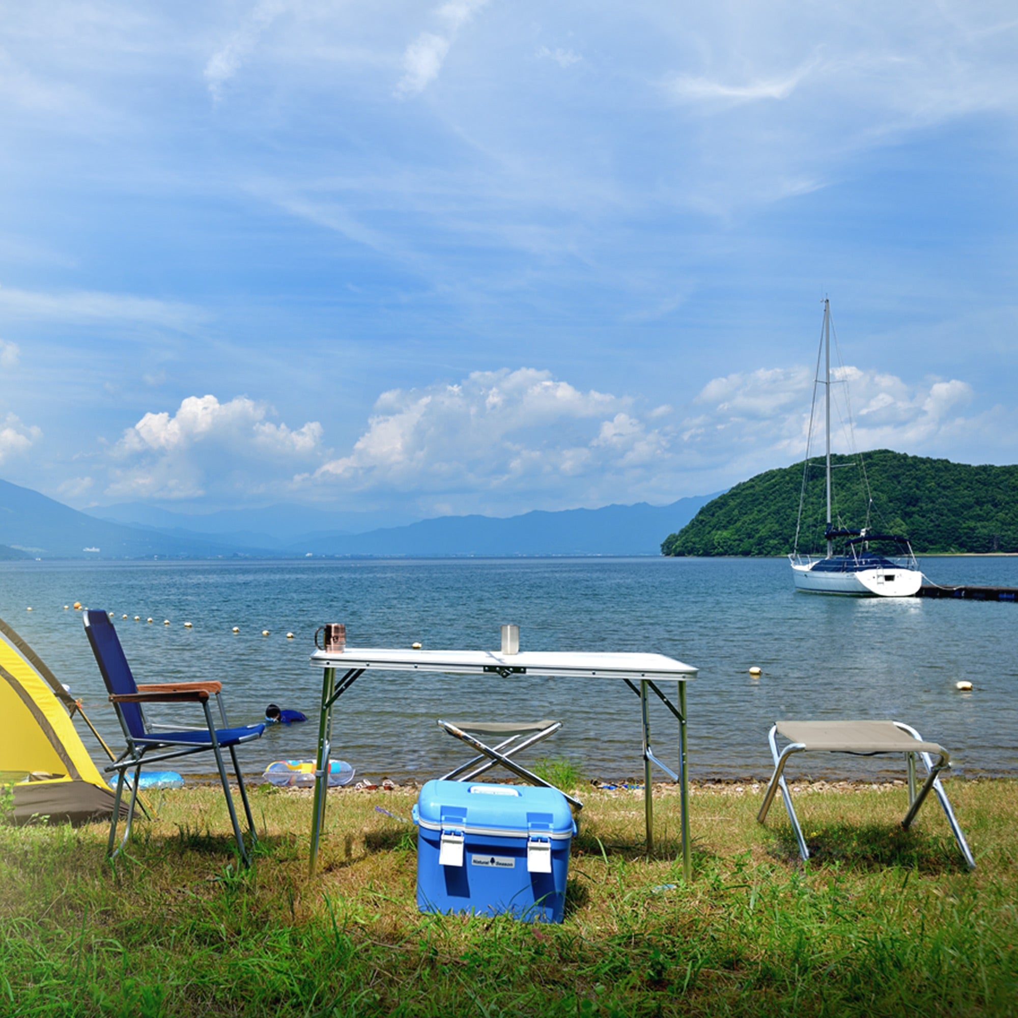 View of a lake with a boat, tables, and chairs on the shore under a blue sky.