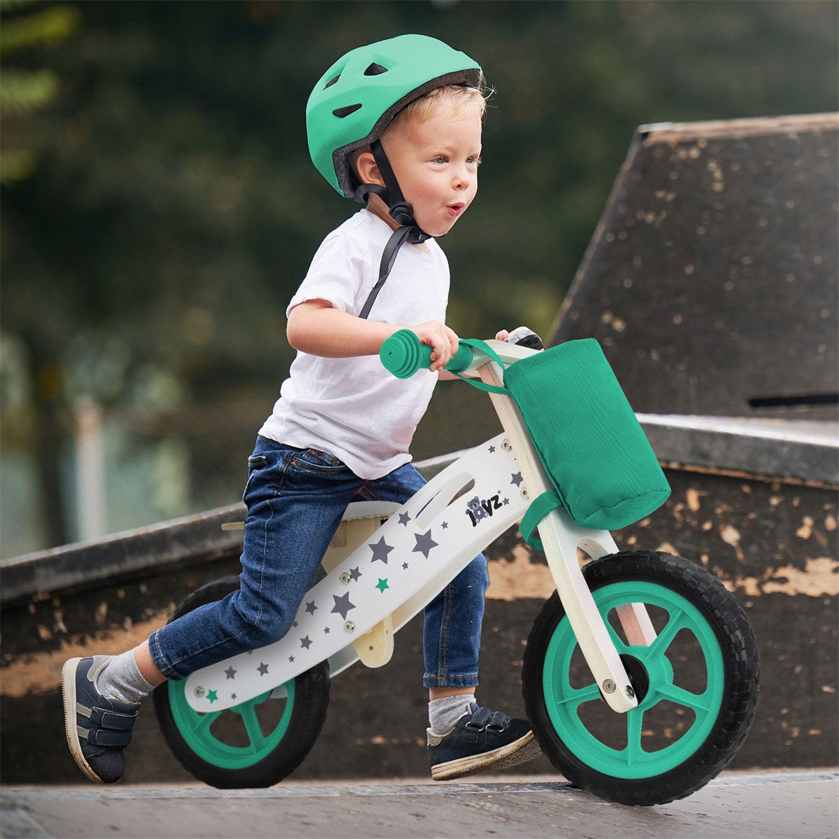 A boy rides a balance bike wearing a green helmet and smiling.