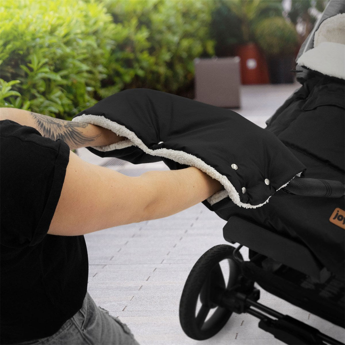 Person holding a black stroller hand muff, with plants in the background.