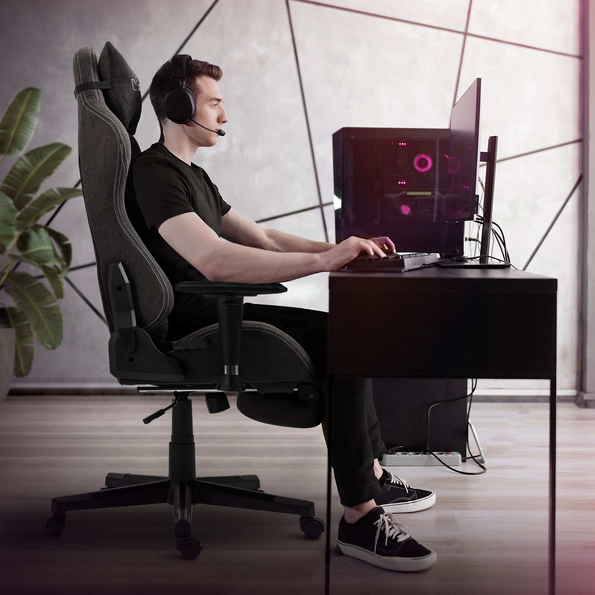 Mann sitzt an Schreibtisch mit Gaming-Stuhl und Computer, trägt Kopfhörer.