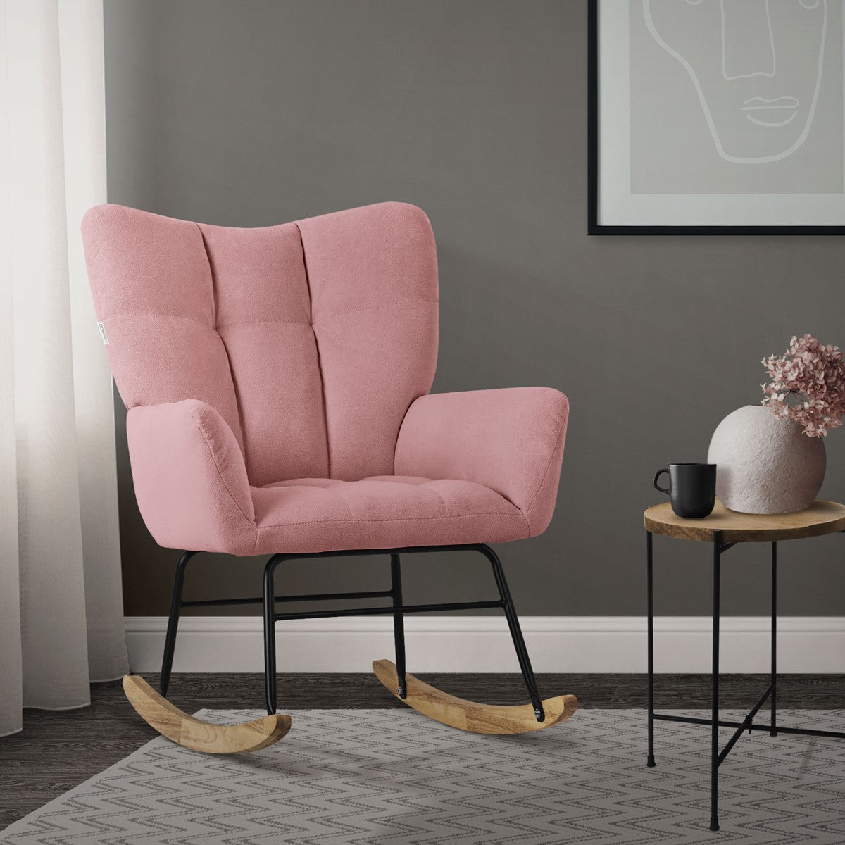 A pink rocking chair stands next to a table with a cup and a vase.