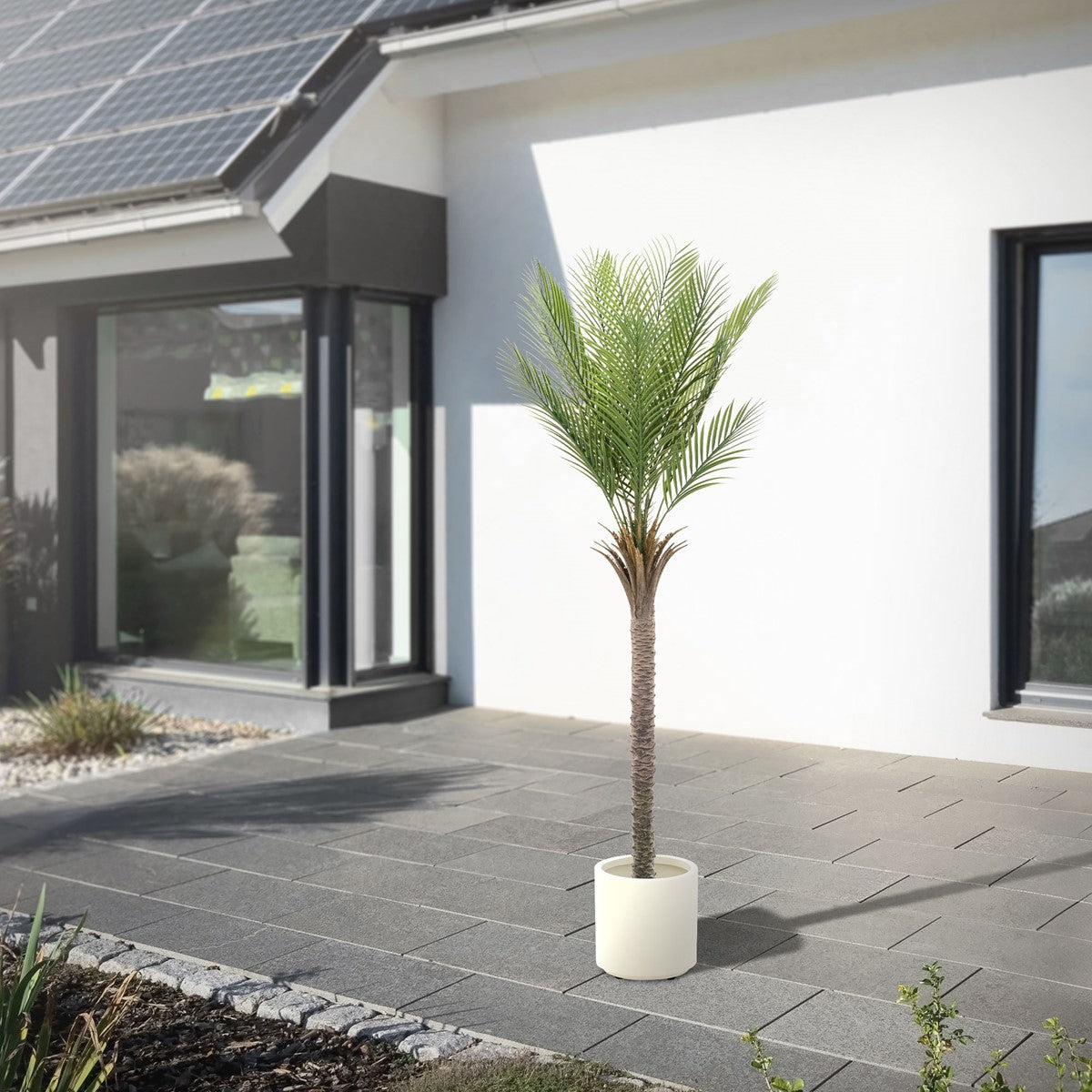 Kunstig palme foran et moderne hus med solpaneler på en terrasse.