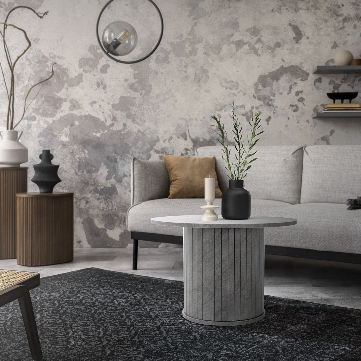 Stue med grå sofa, rundt bord og dekorative planter på et tæppe.
