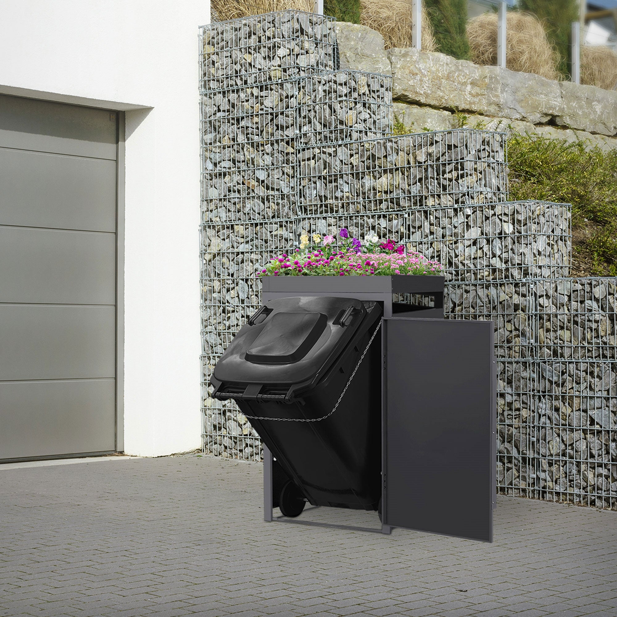Mülltonnenbox mit Blumenbepflanzung, auf einem gepflasterten Weg.