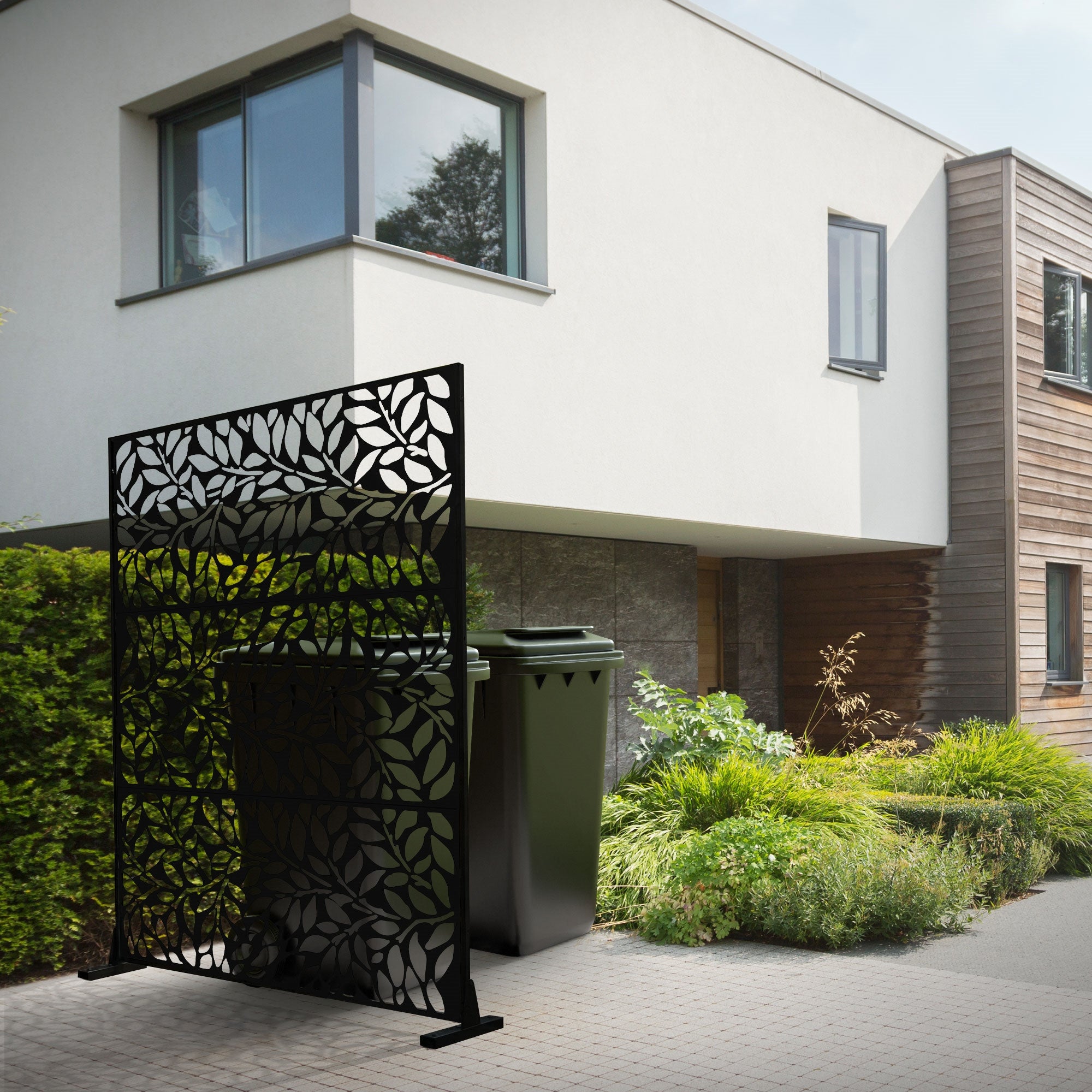 Sort skærmvæg med bladmønster foran en moderne husfacade og affaldsbeholdere.