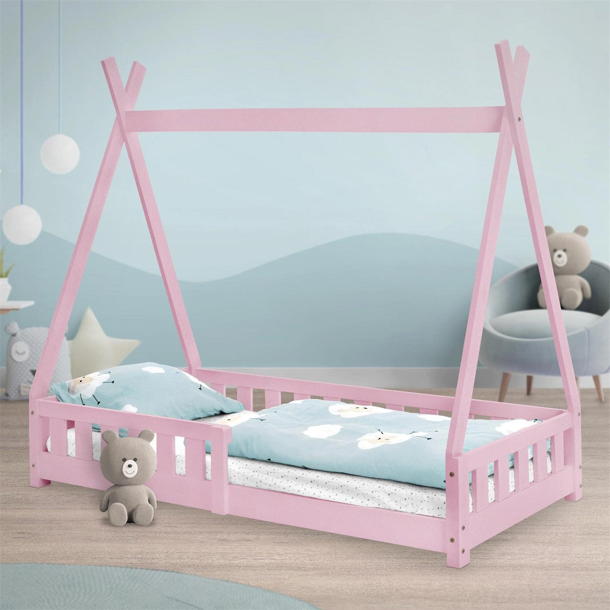 Een roze kinderbed met kussens en een teddybeer op een houten vloer.