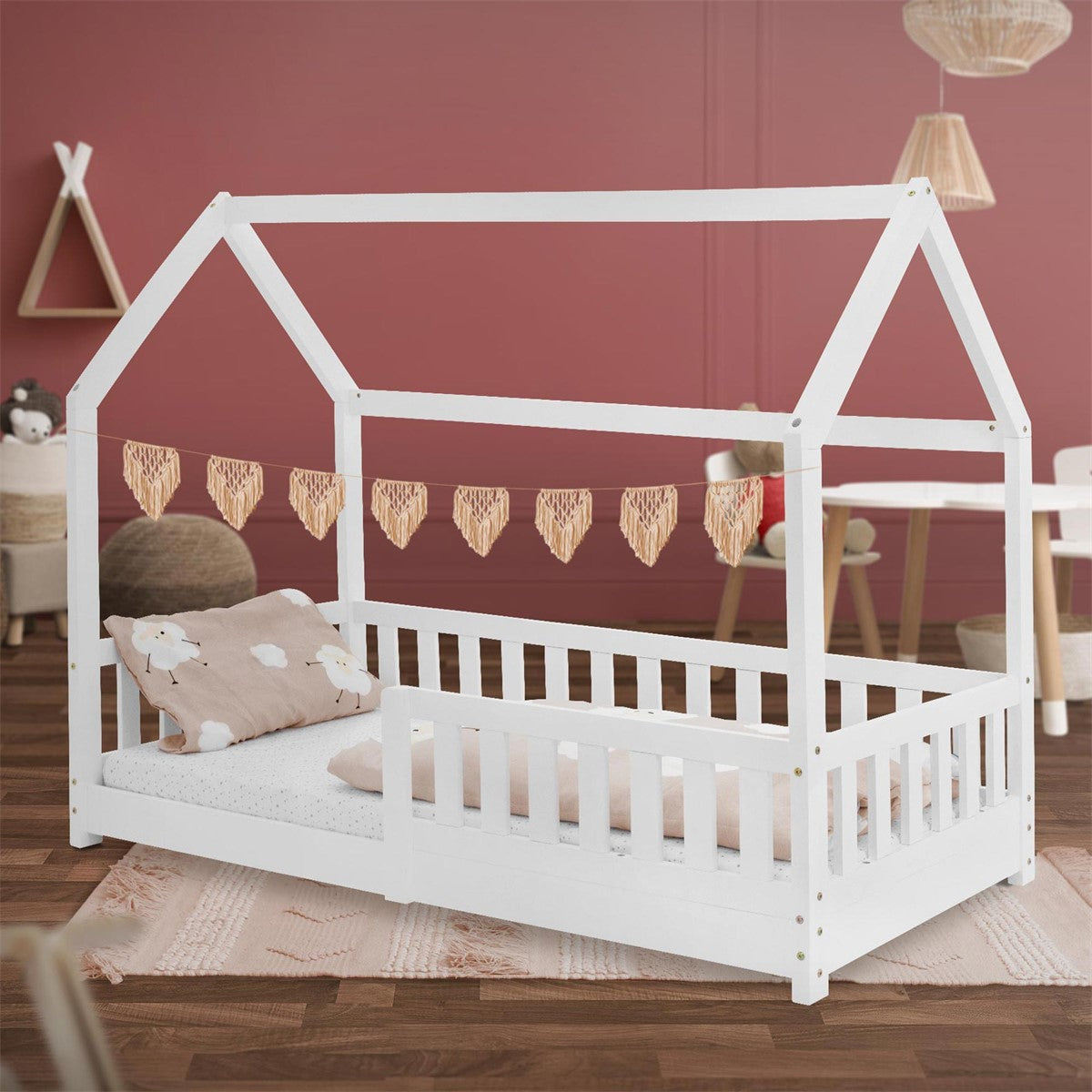 Weißes Kinderbett im Hausdesign mit Kissen und einer Girlande aus Holzherzen.
