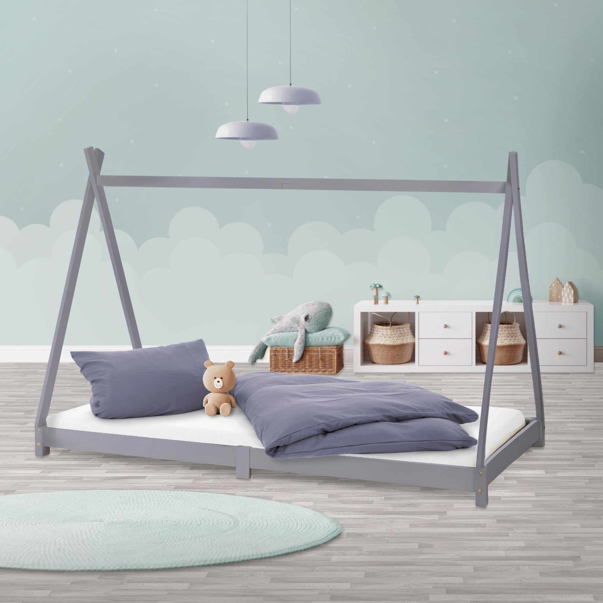 Ein hellgraues Kinderbett mit Kissen und Decke, einem Teddy und einem hellen Hintergrund.