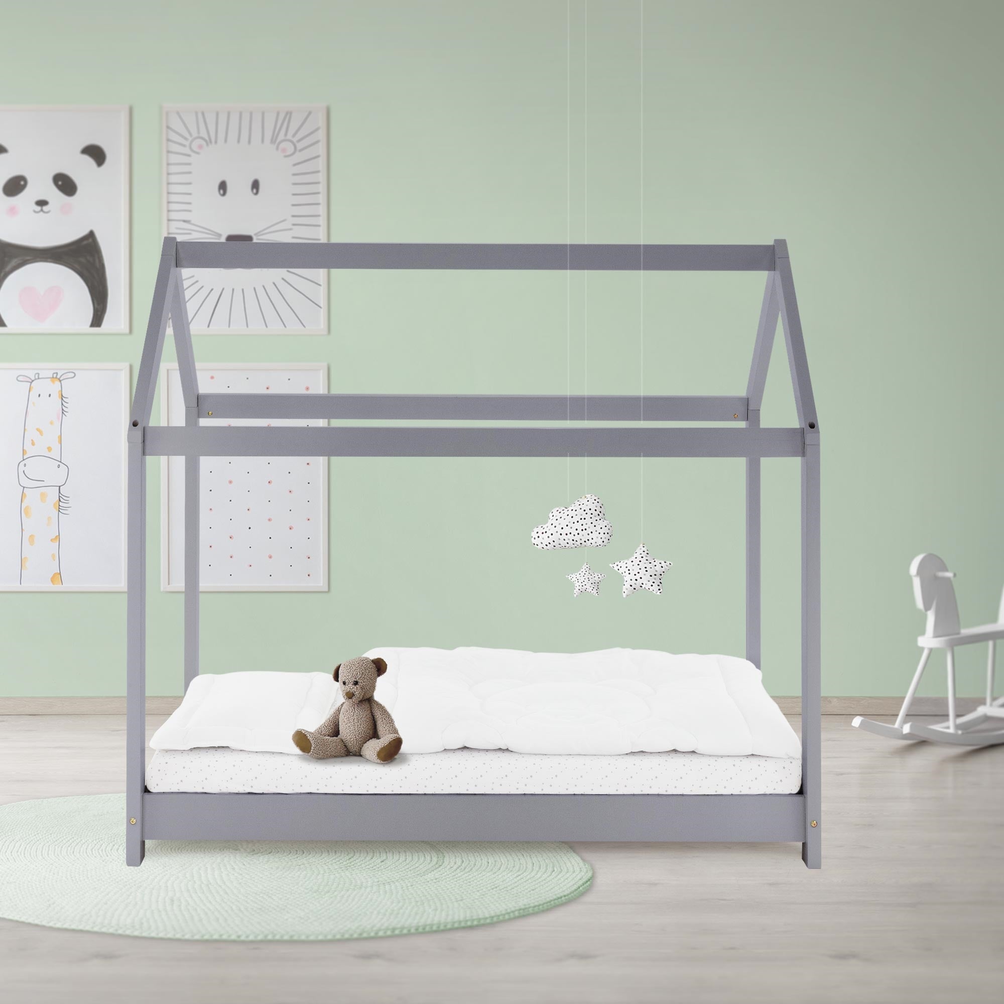 Ein Kinderbett in einem hellgrünen Raum mit Bildern an der Wand und einem Teddy auf dem Bett.