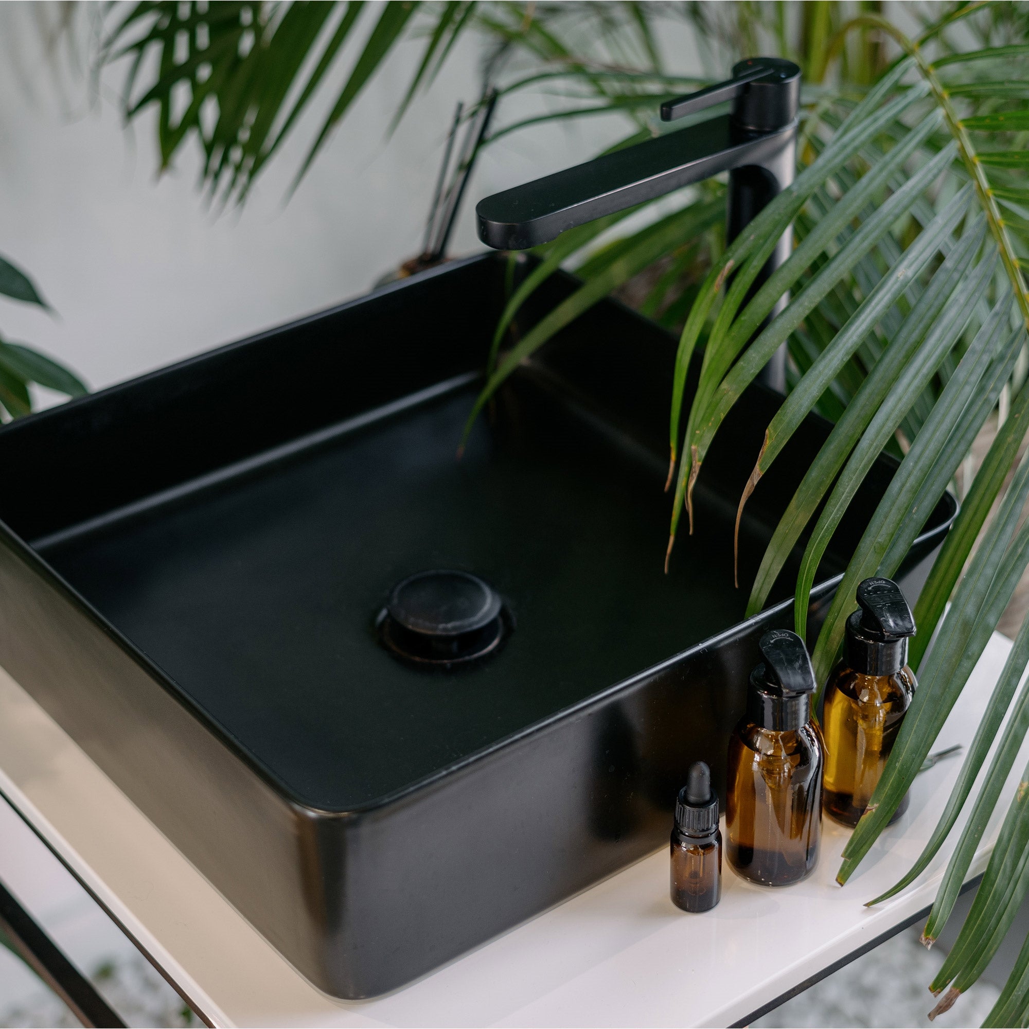 Black sink surrounded by plants and bottles on a table.