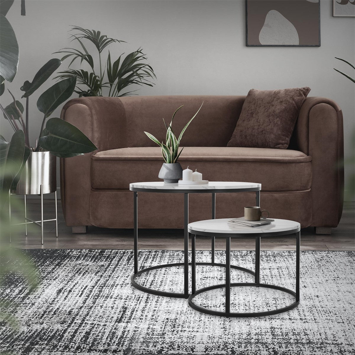 Sala de estar com um sofá castanho, duas mesas e plantas ao fundo.