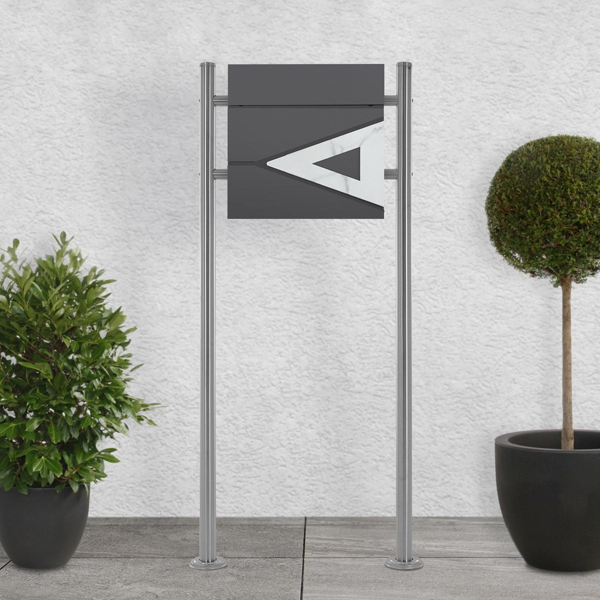 Briefkasten mit grauer Front und Holzdetails, auf einem Gehweg neben Pflanzen.