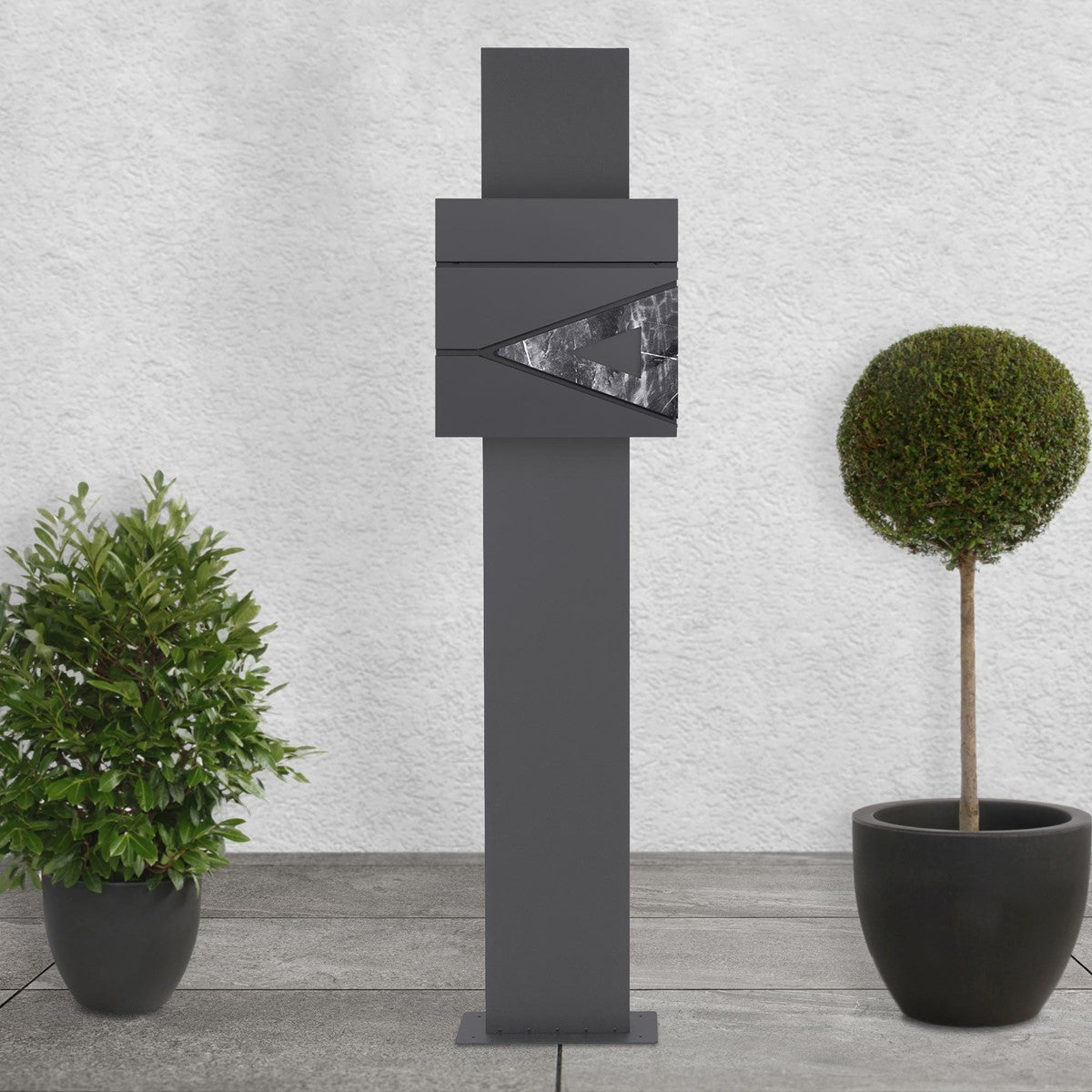 Anthracite mailbox with a marble look, surrounded by plants on a walkway.