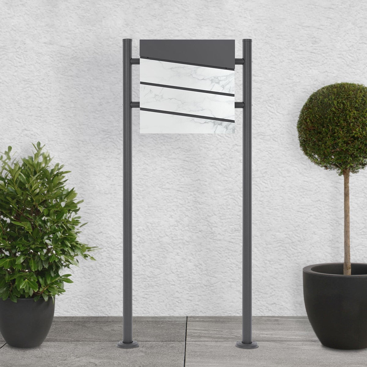Letterbox with a grey lid and three white fronts, surrounded by plants.