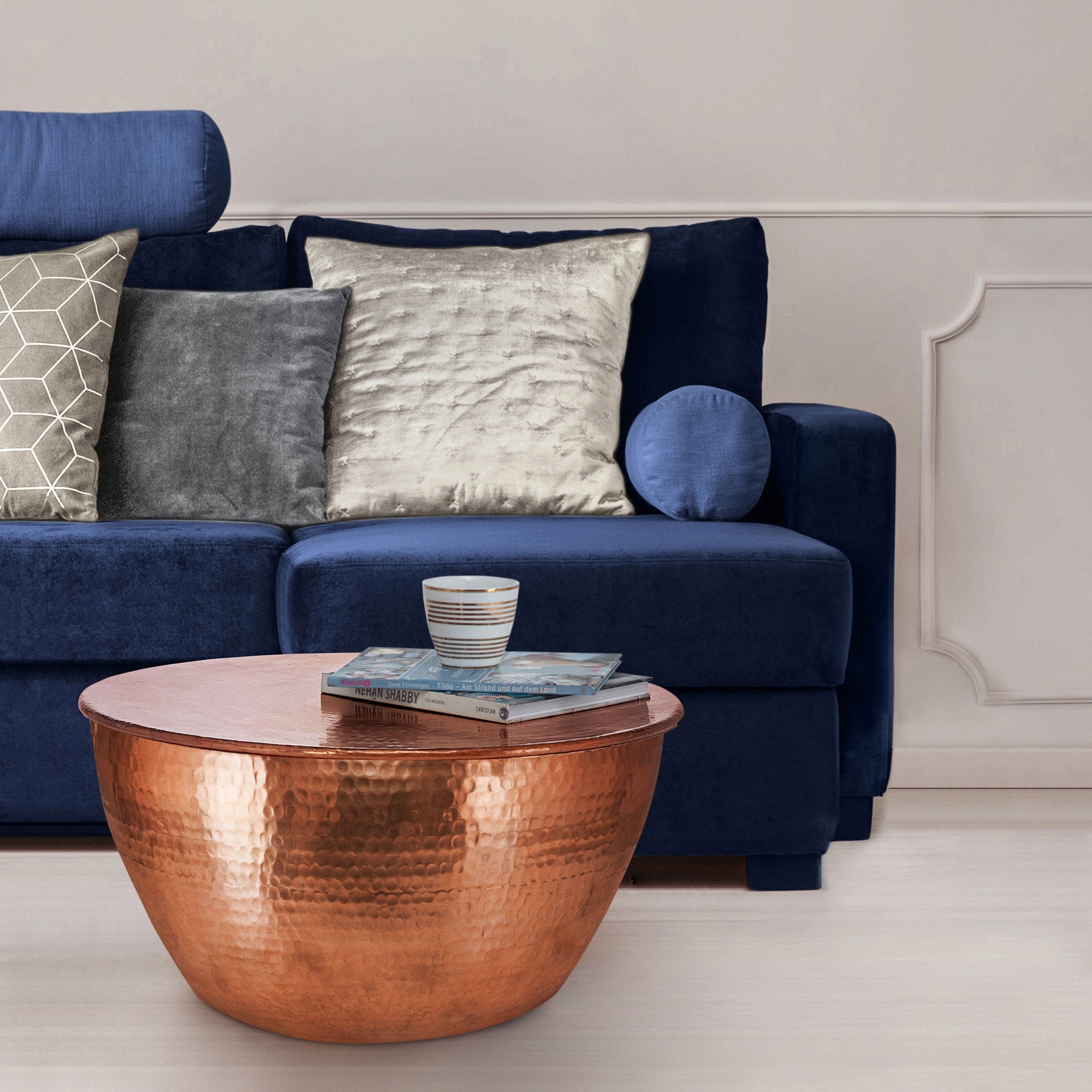 Blaues Sofa mit Kissen und einem runden Kupfertisch, darauf Bücher und ein Glas.