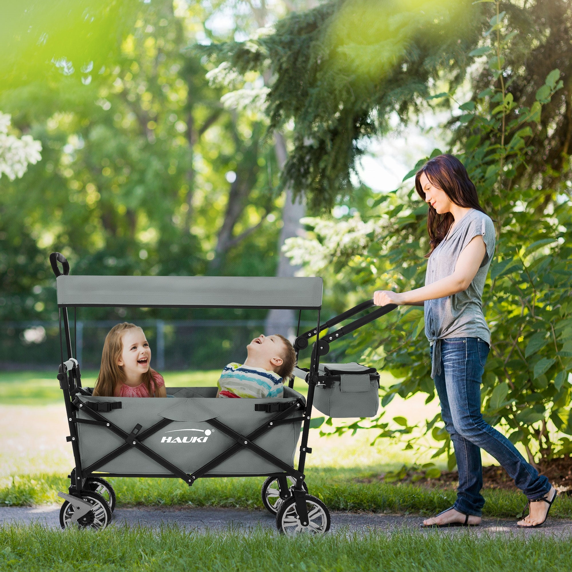 Eine Frau schiebt einen grauen Kinderwagen mit zwei Kindern im Park.