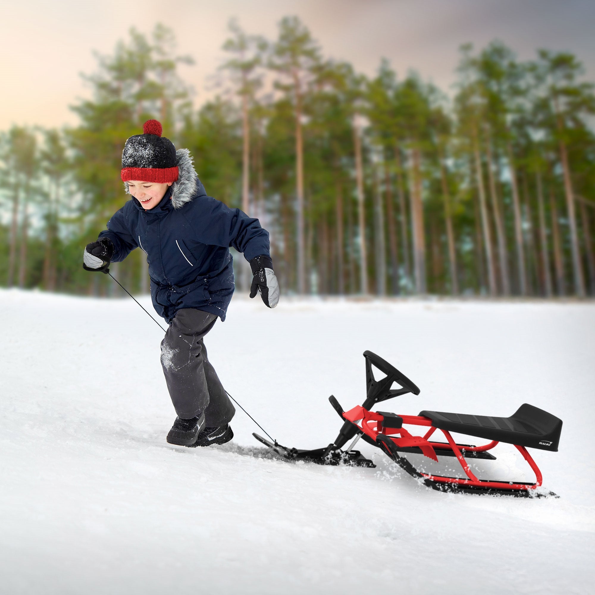 Child pulling a red steering sled in the snow, surrounded by trees.