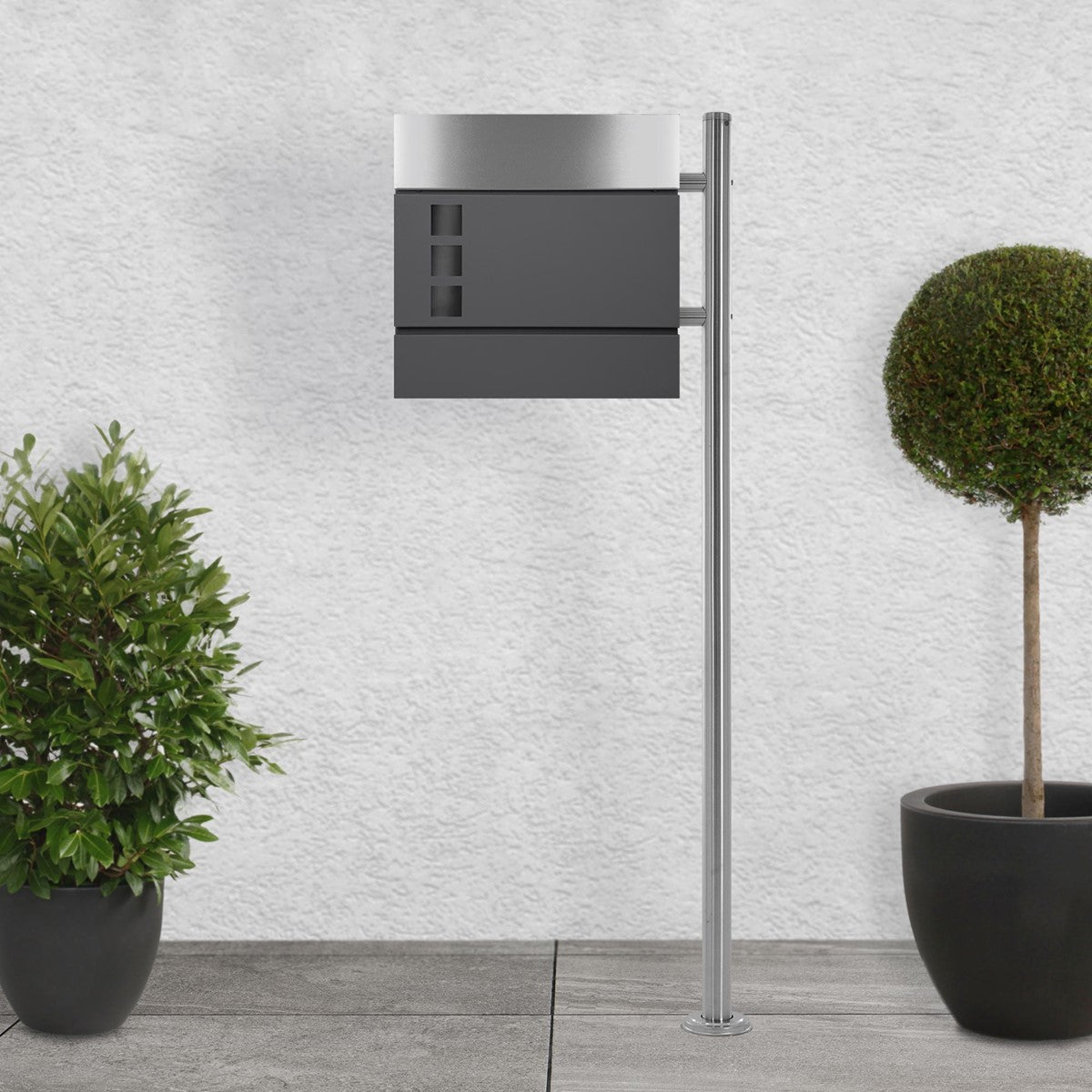 Freestanding mailbox with a curved front and two plants in the background