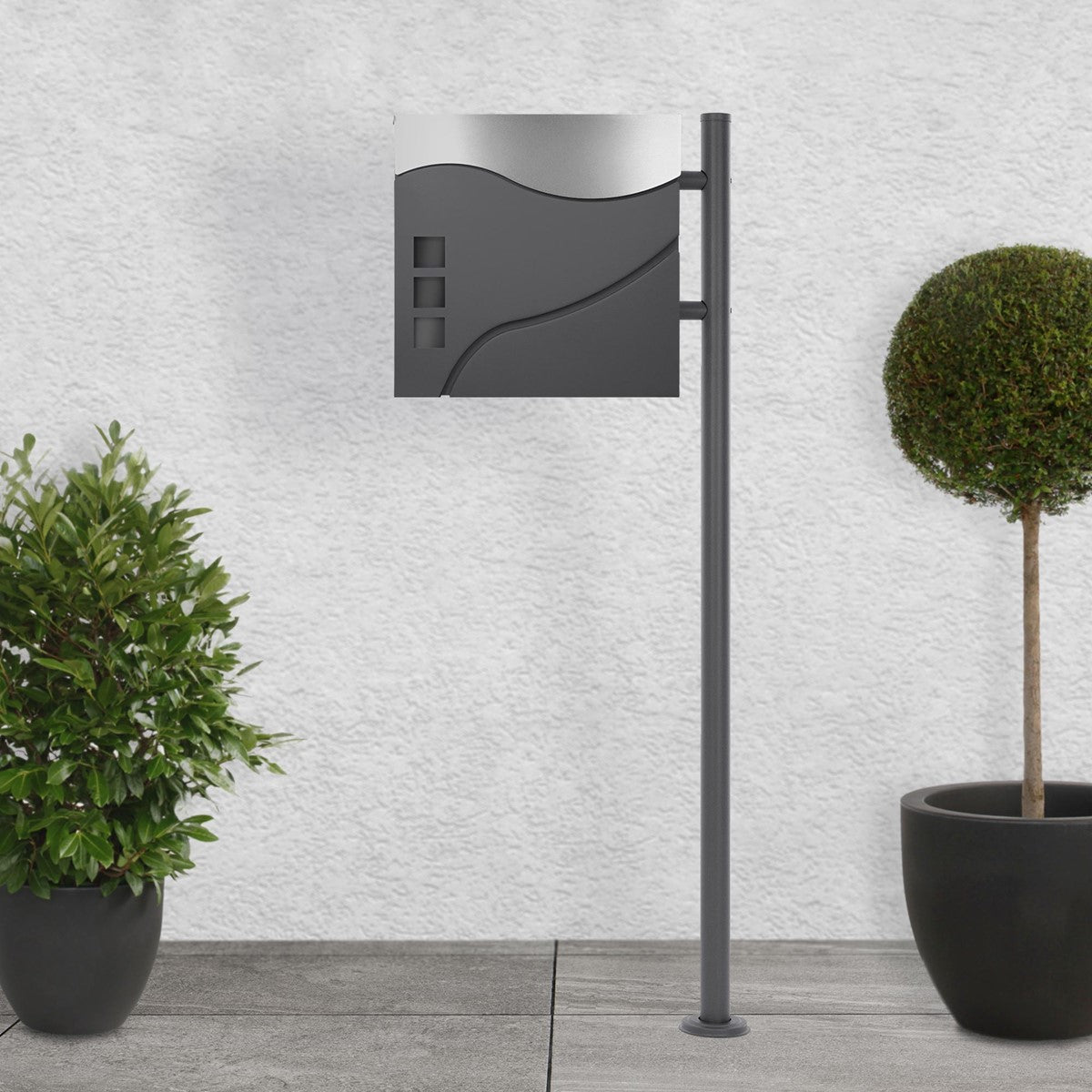 Freestanding mailbox with a curved front and two plants in the background
