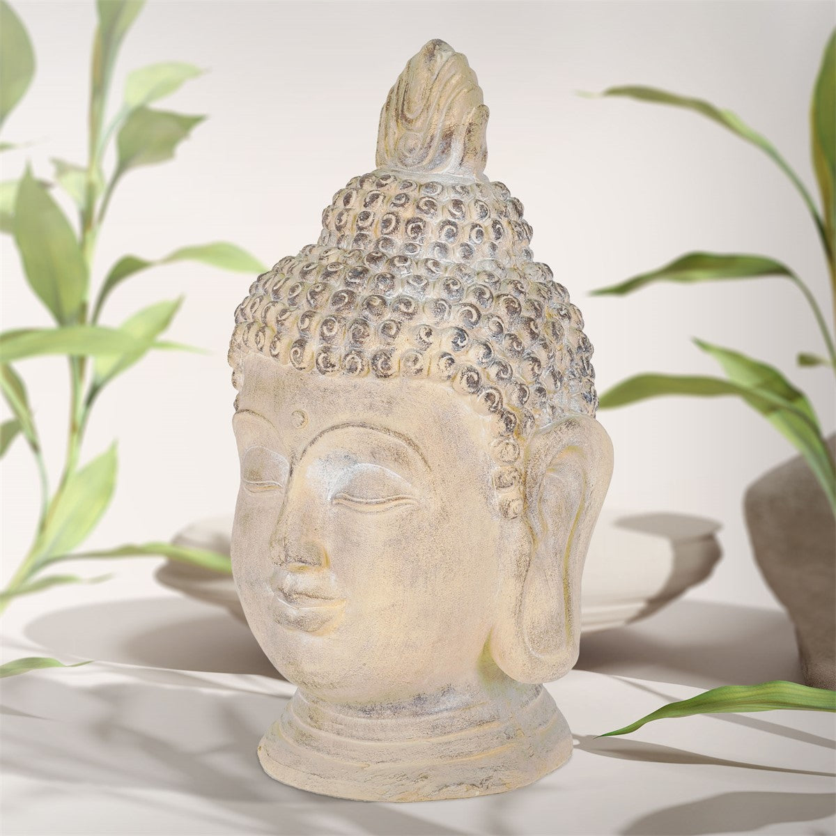 Buddha-Kopf aus Kunstharz, seitliche Ansicht, mit dekorativen Details und Pflanzen im Hintergrund