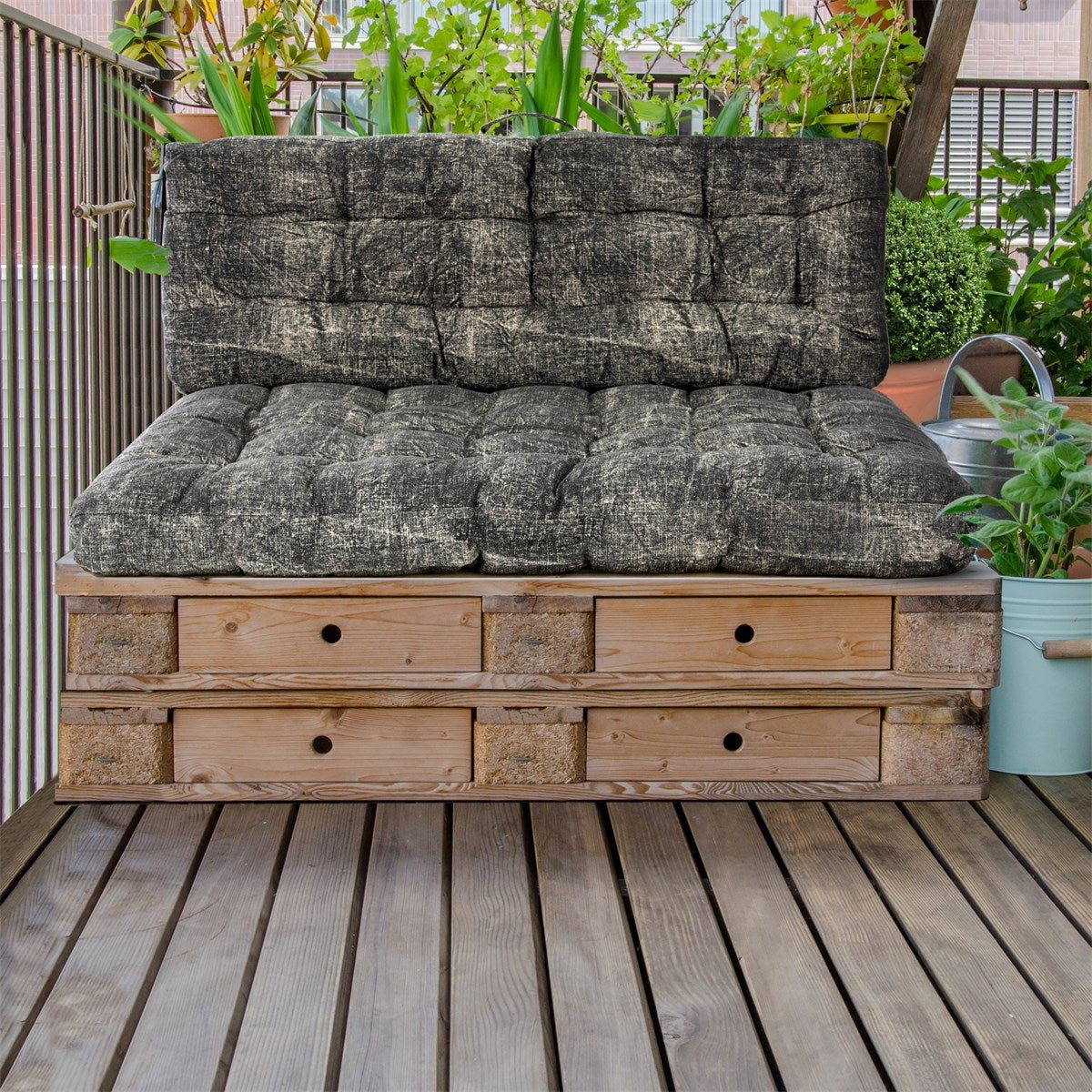Seduta realizzata con pallet di legno, con cuscini grigi e piante sullo sfondo.