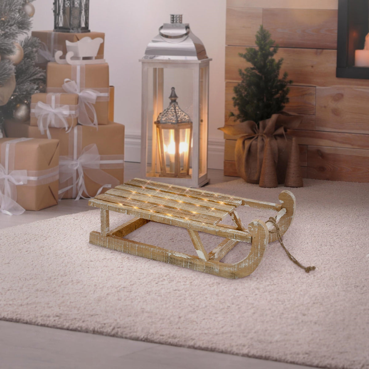 Ein Holzschlitten steht auf einem Teppich vor einer Laterne und einem Weihnachtsbaum.