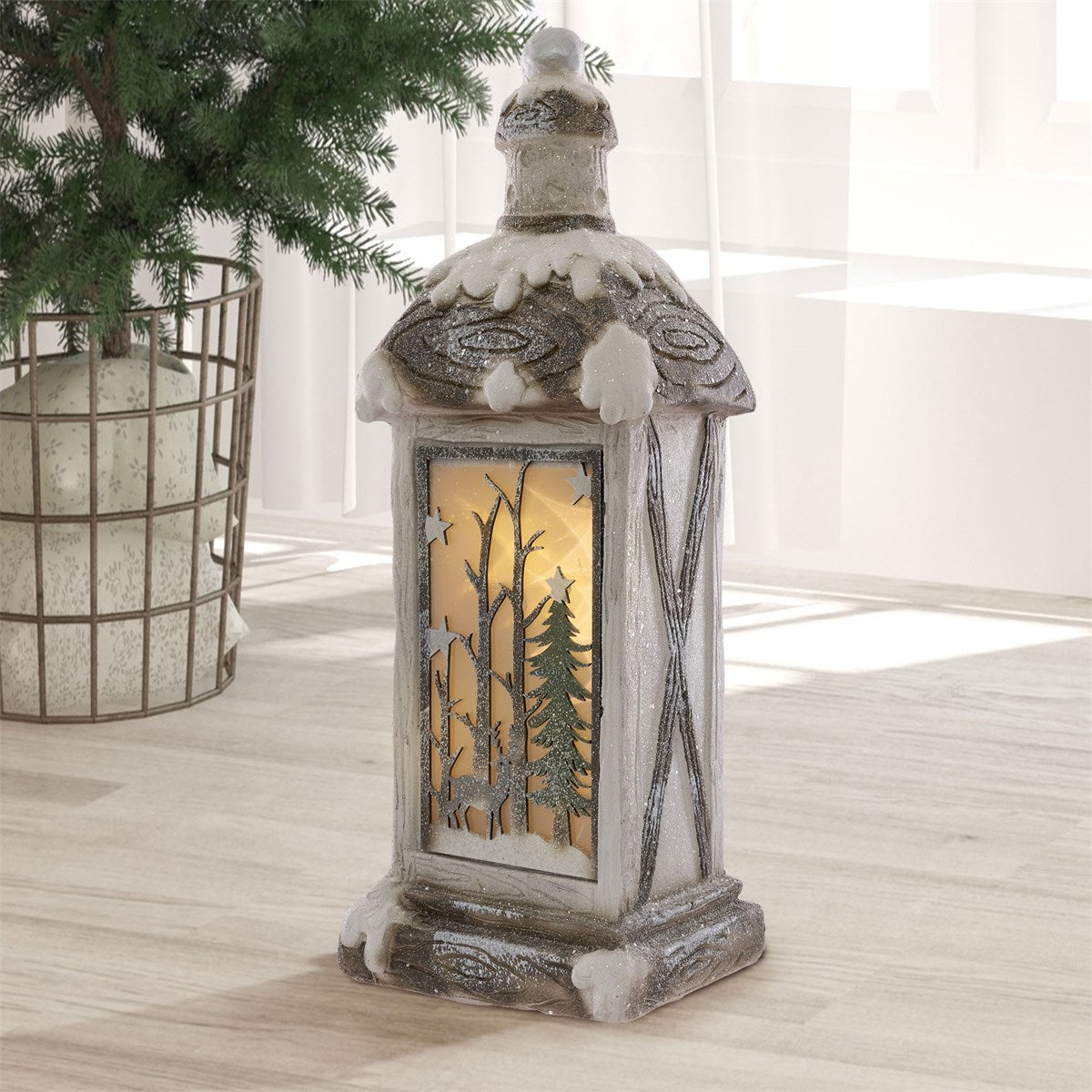 Lanterna decorativa con tetto innevato e finestra illuminata con motivi di abeti e stelle, su una superficie chiara.
