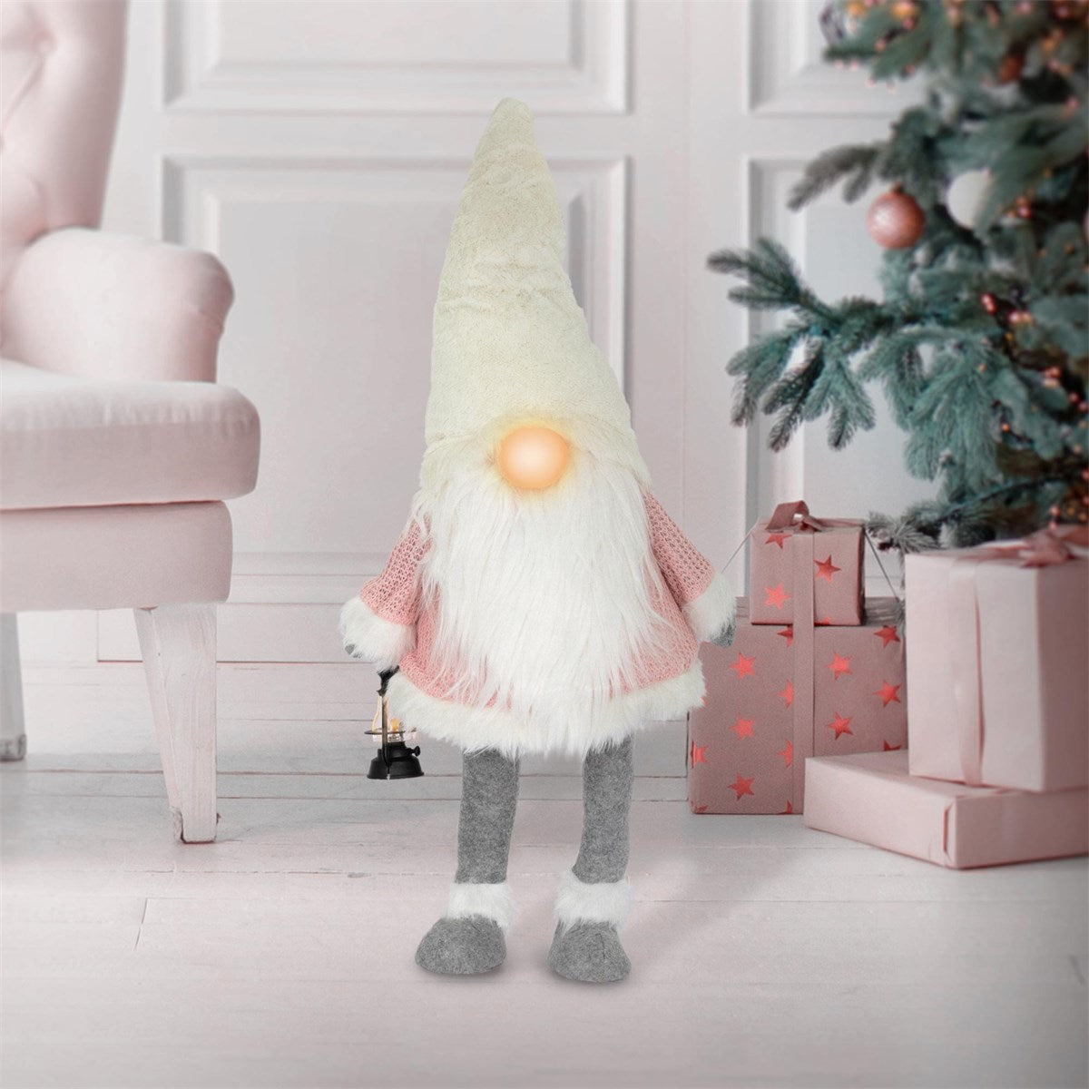 Ein dekorativer Wichtel mit weißem Bart und einer Glocke, vor einem Weihnachtsbaum und Geschenken