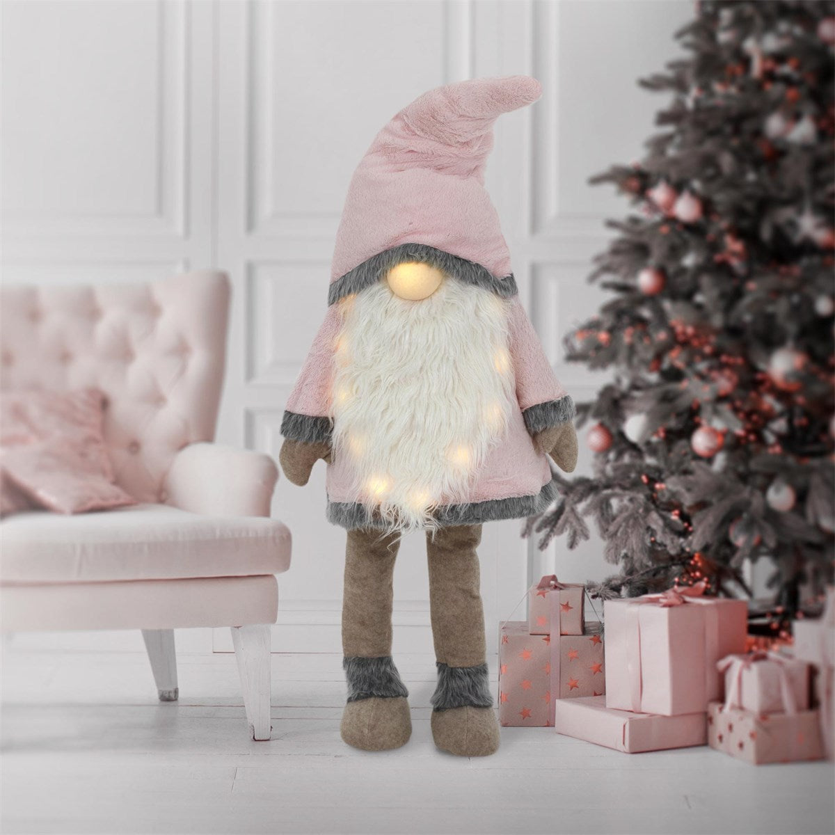 Lo gnomo di stoffa rosa è in piedi davanti a un albero di Natale e a dei regali, con luci sul corpo.